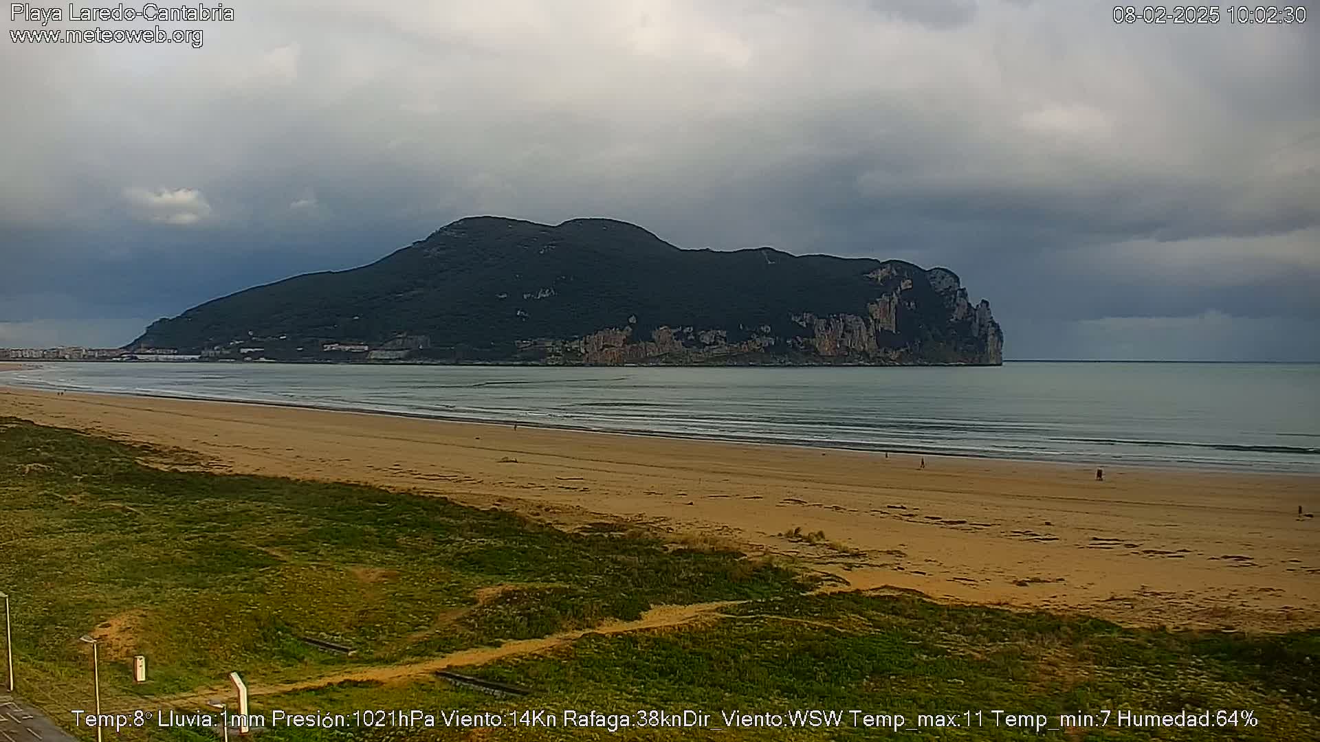 A sandy beach stretches out to a calm sea under an overcast sky, with a large, dark, mountainous island visible in the distance.
