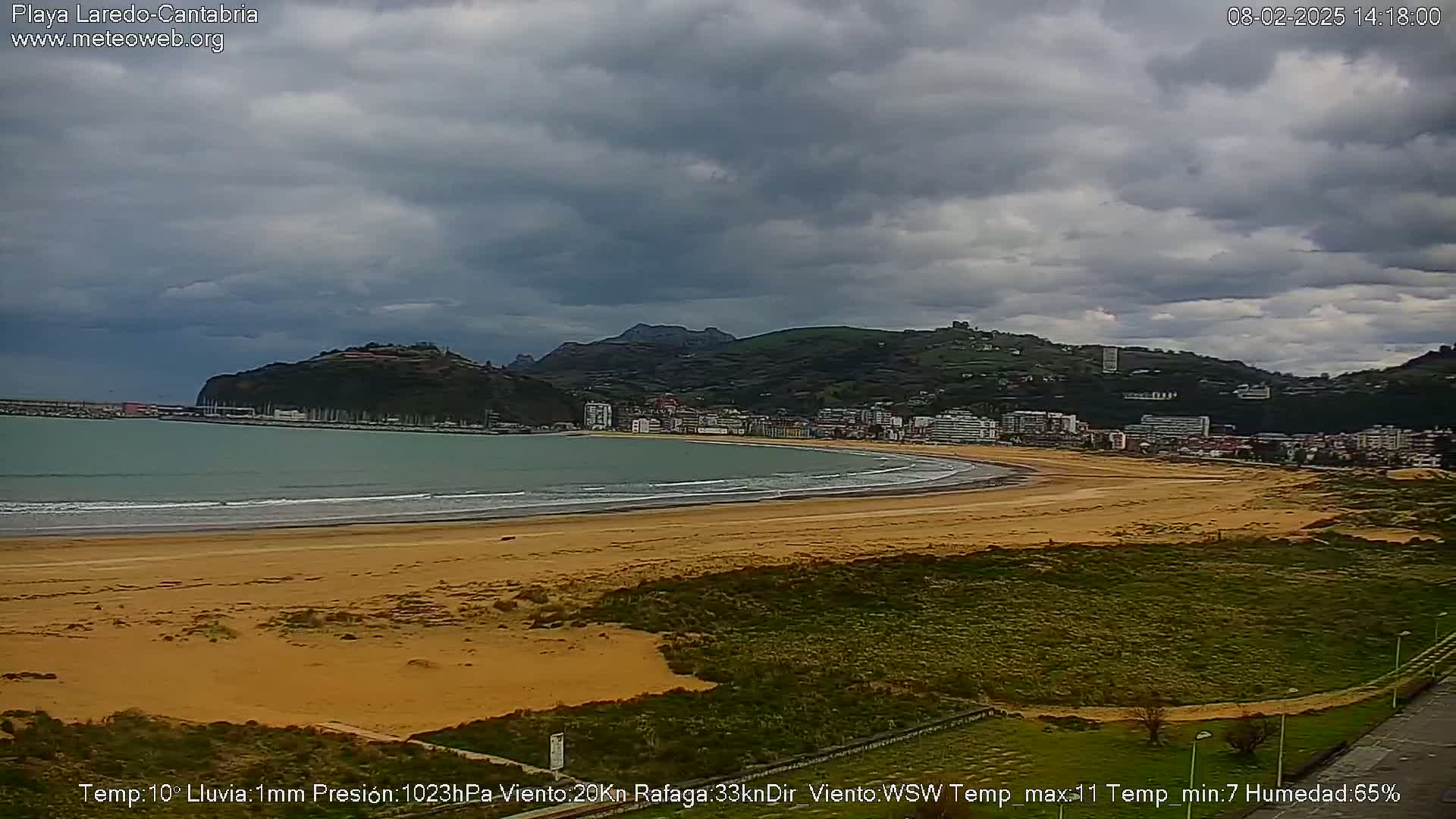 A long sandy beach curves along a coastline under overcast skies, backed by a town nestled against green hills.