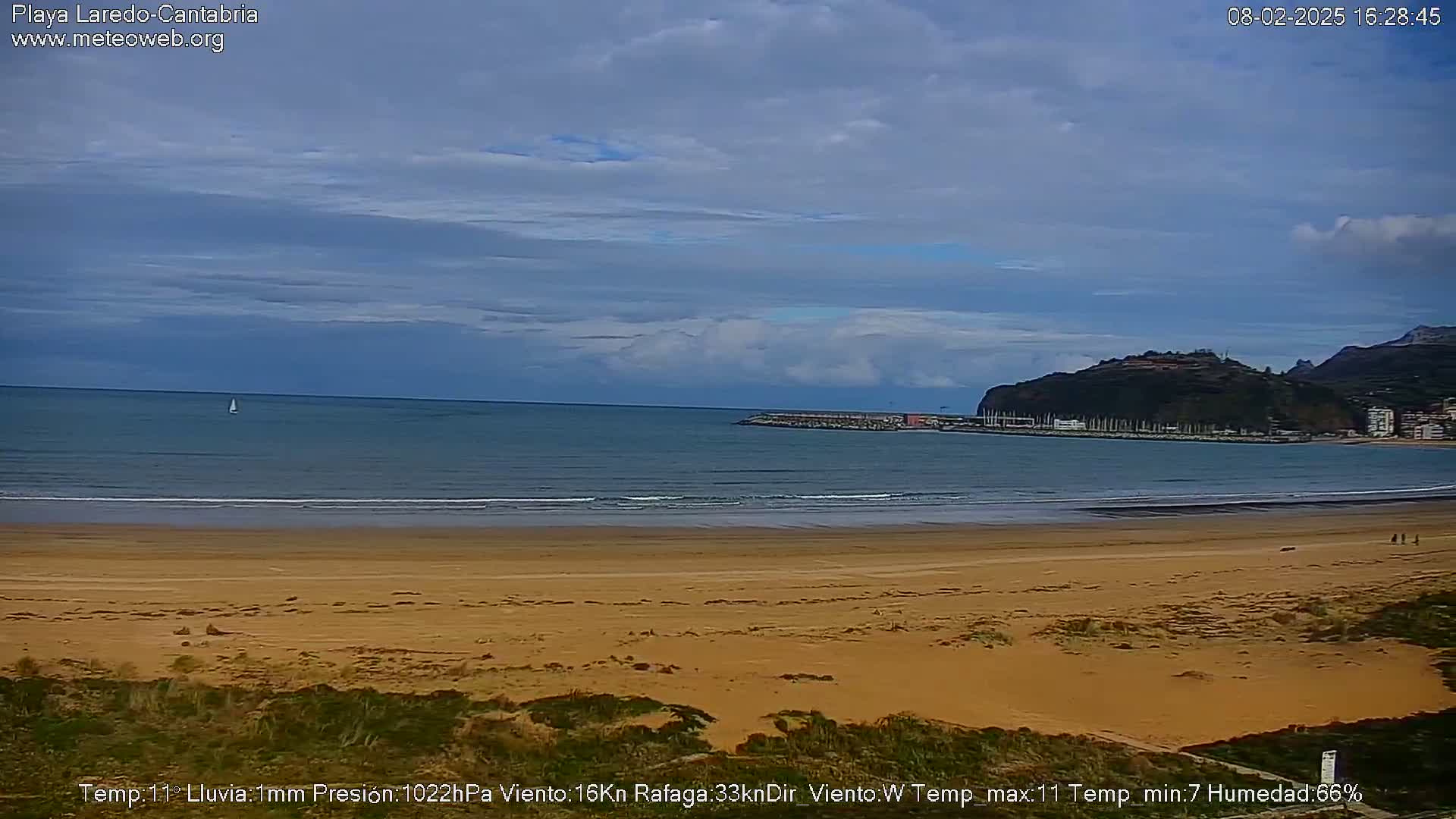 A sandy beach stretches towards a calm ocean under a mostly cloudy sky, with a distant hill visible in the background.