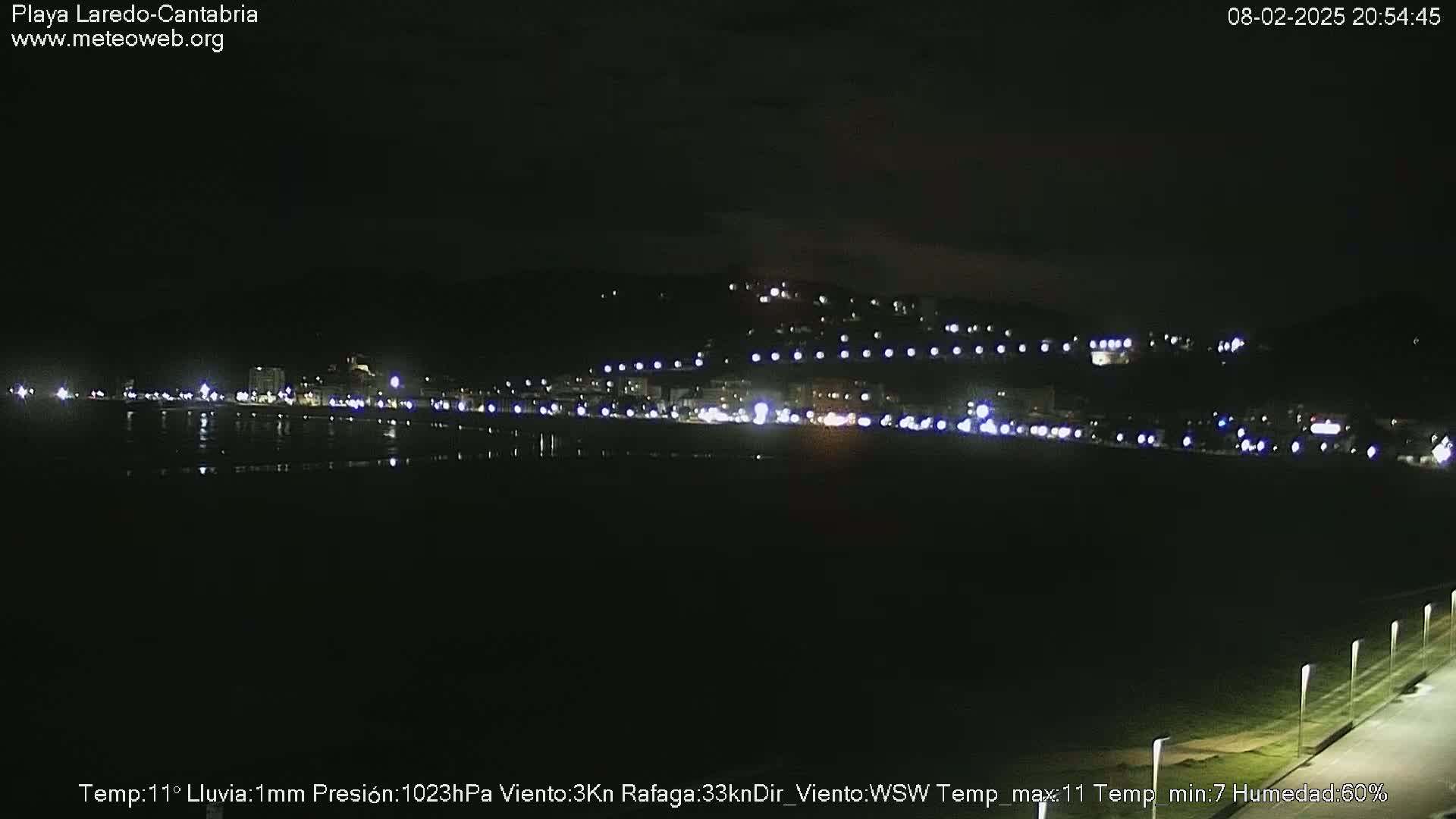 A nighttime view of a dark beach and a coastal town illuminated by lights, under a dark sky with light rain.