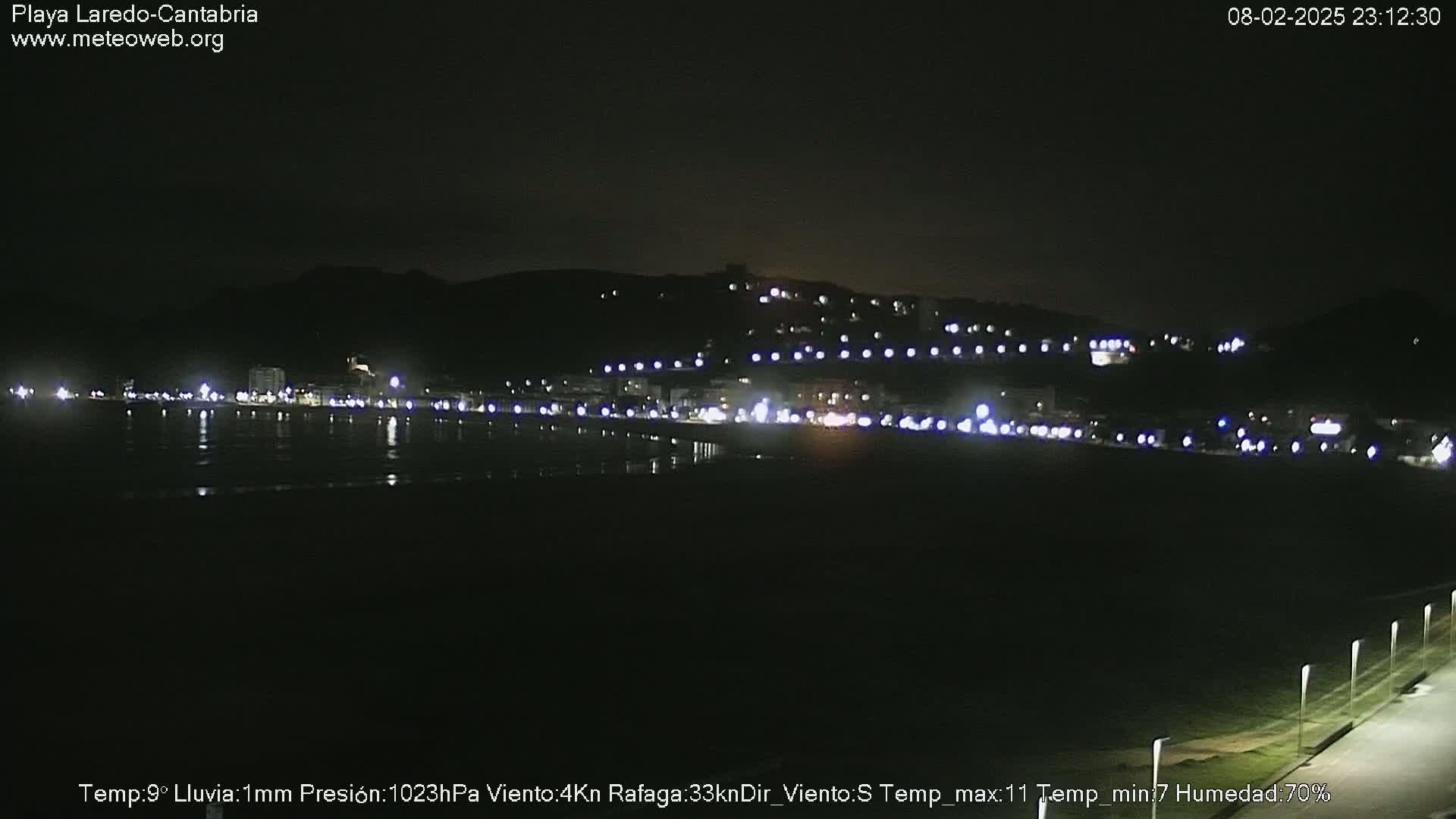 A nighttime view of a dark beach and illuminated town on a hillside, with light rain.