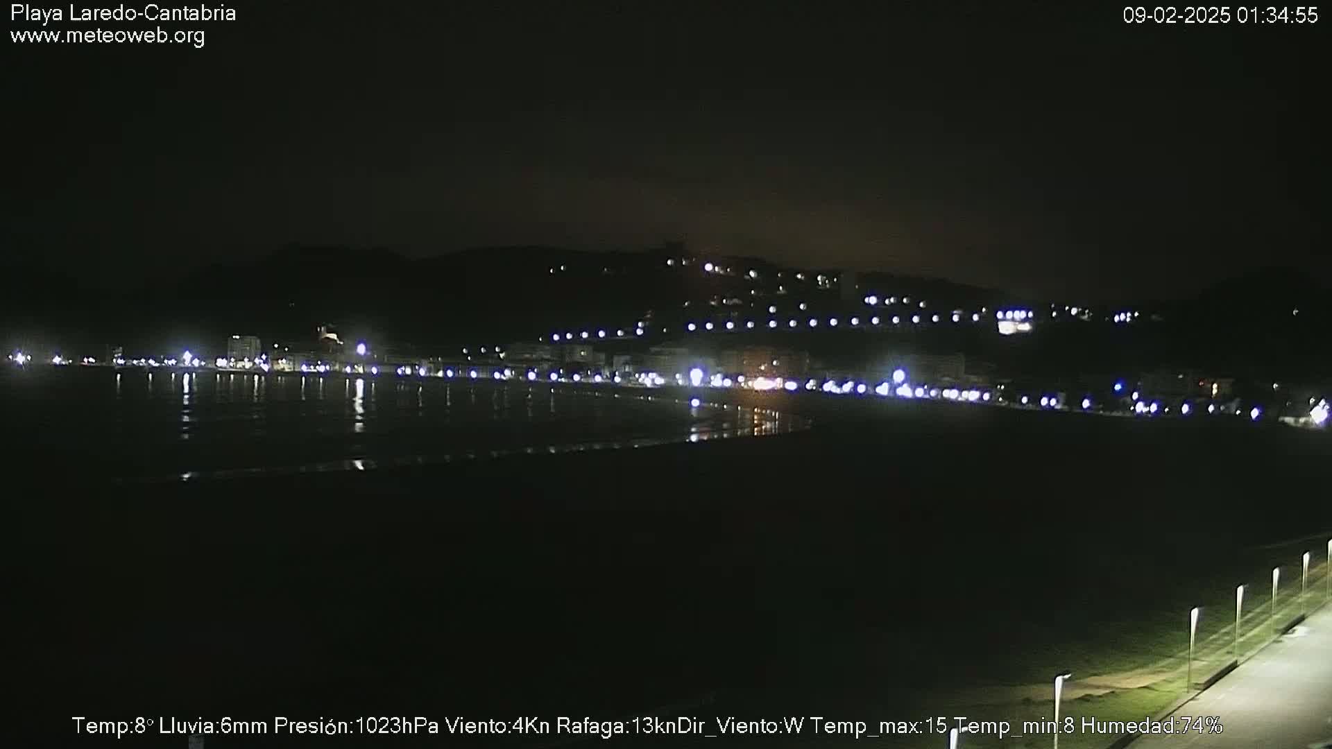 A nighttime view of a dark beach with a town's lights illuminating the coast under a clear sky, with light rain.