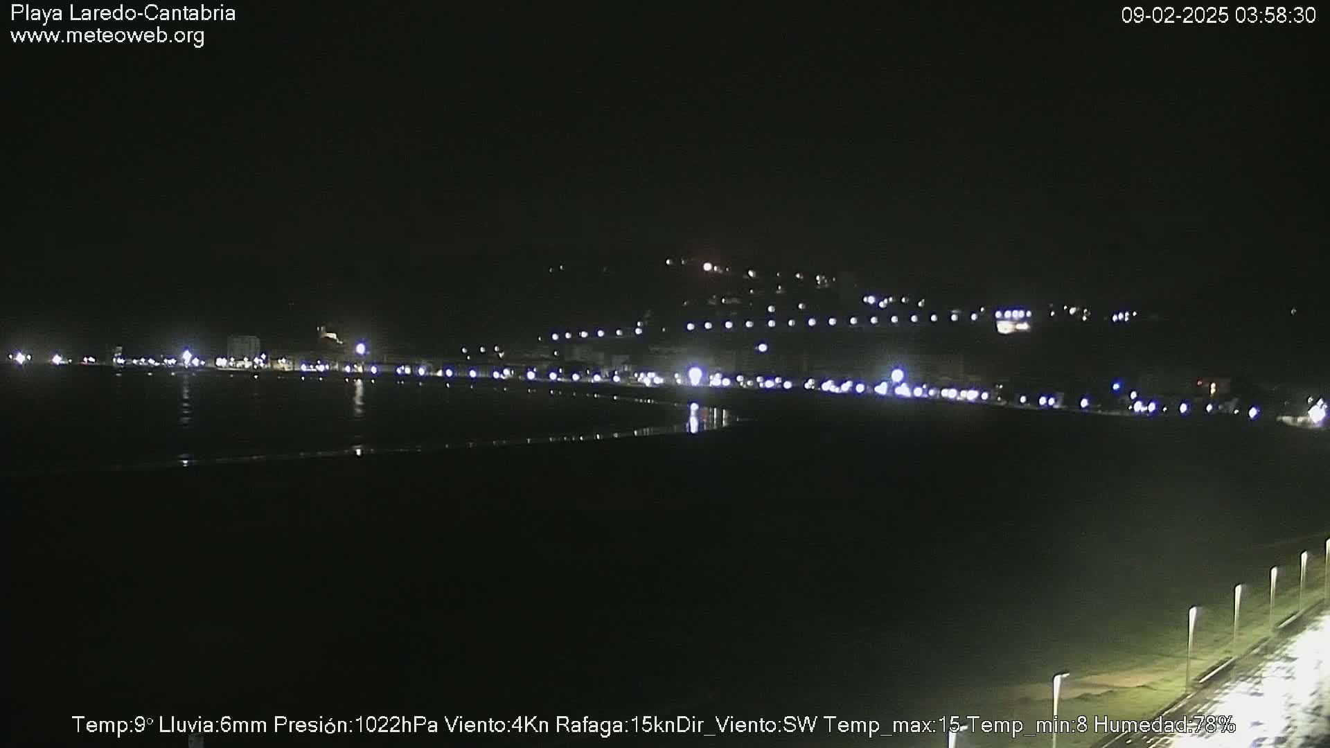 A nighttime view of a dark beach with a line of lights along the shore and hills in the background, under conditions of rain.