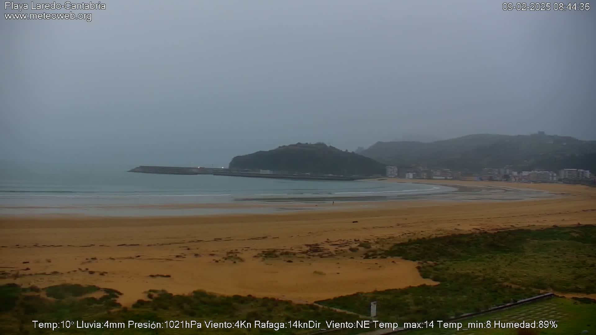 A wide shot of a sandy beach and calm ocean under an overcast sky, with a hill and town visible in the distance.