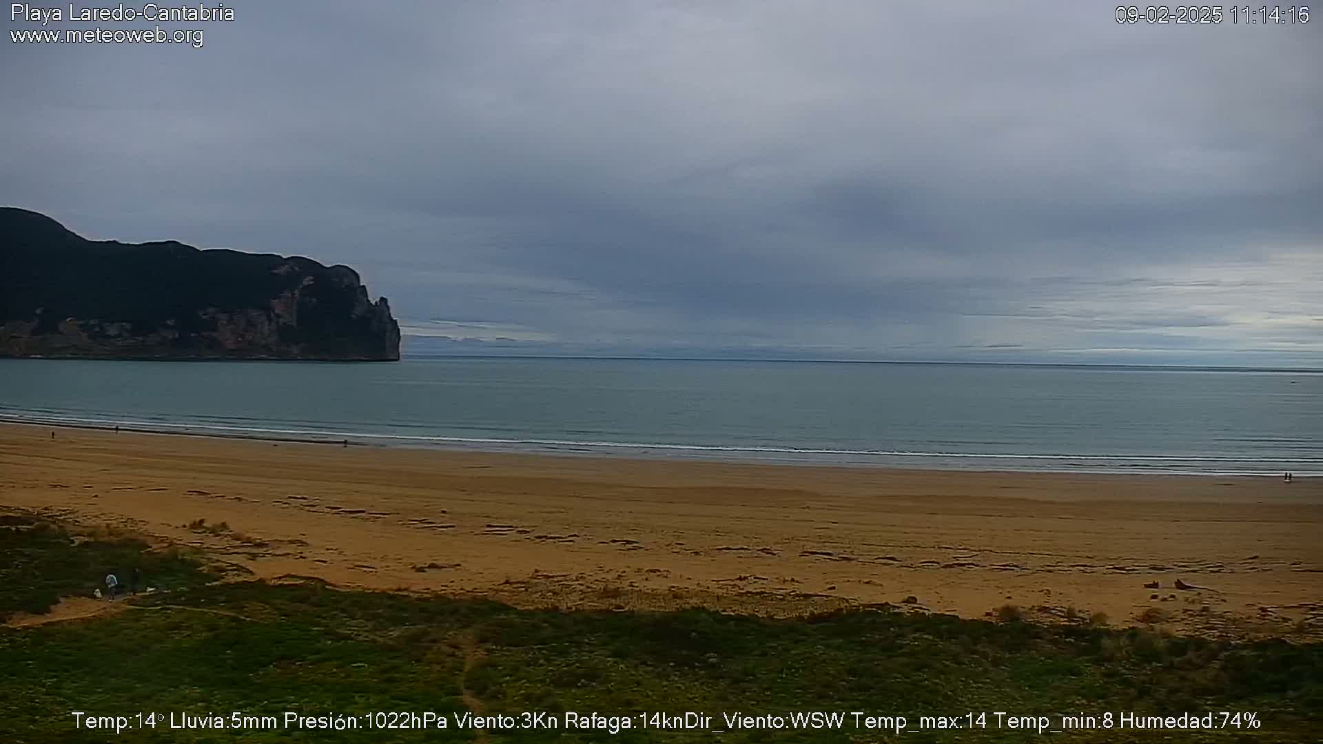 A wide sandy beach stretches to a calm, gray-toned ocean under an overcast sky, with a dark, rocky headland visible in the distance.