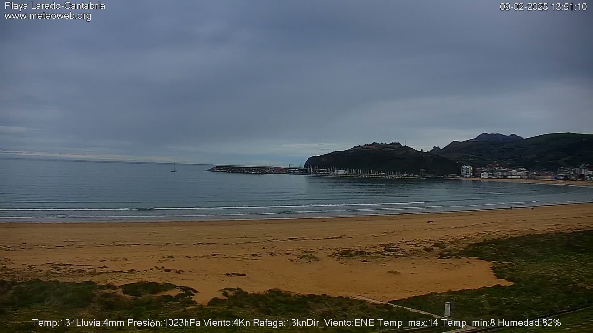 A sandy beach stretches along a calm ocean under an overcast sky, with a small town nestled against a hill in the background.