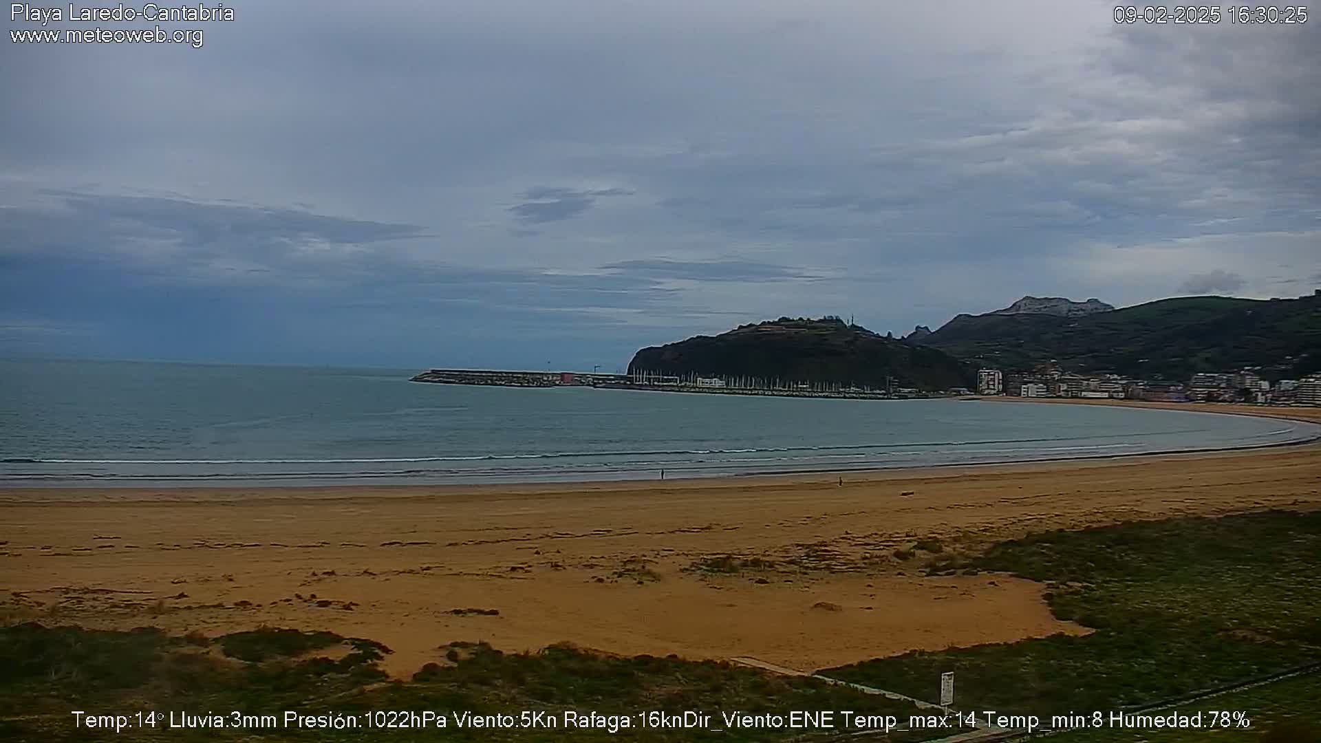A sandy beach curves around a calm bay under an overcast sky, with hills and a town visible in the distance.