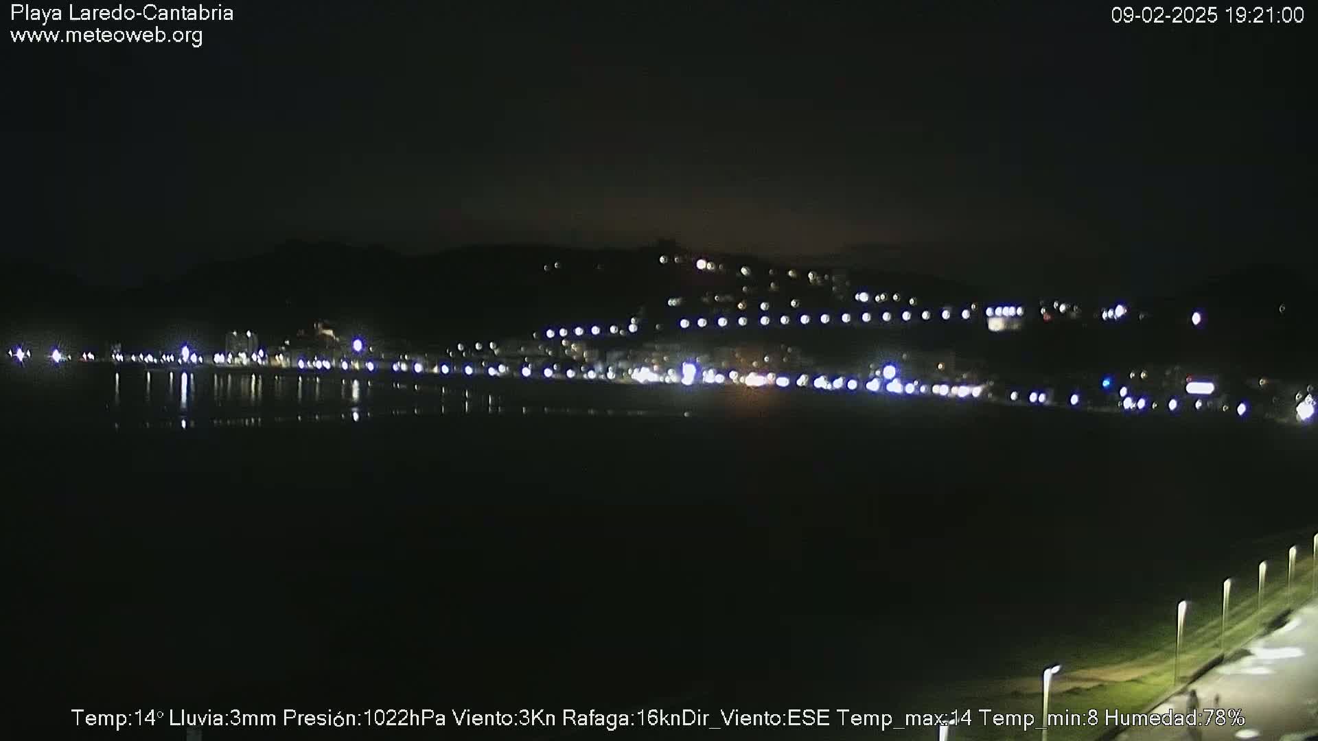 A nighttime view of a dimly lit town beside a dark body of water under light rain.