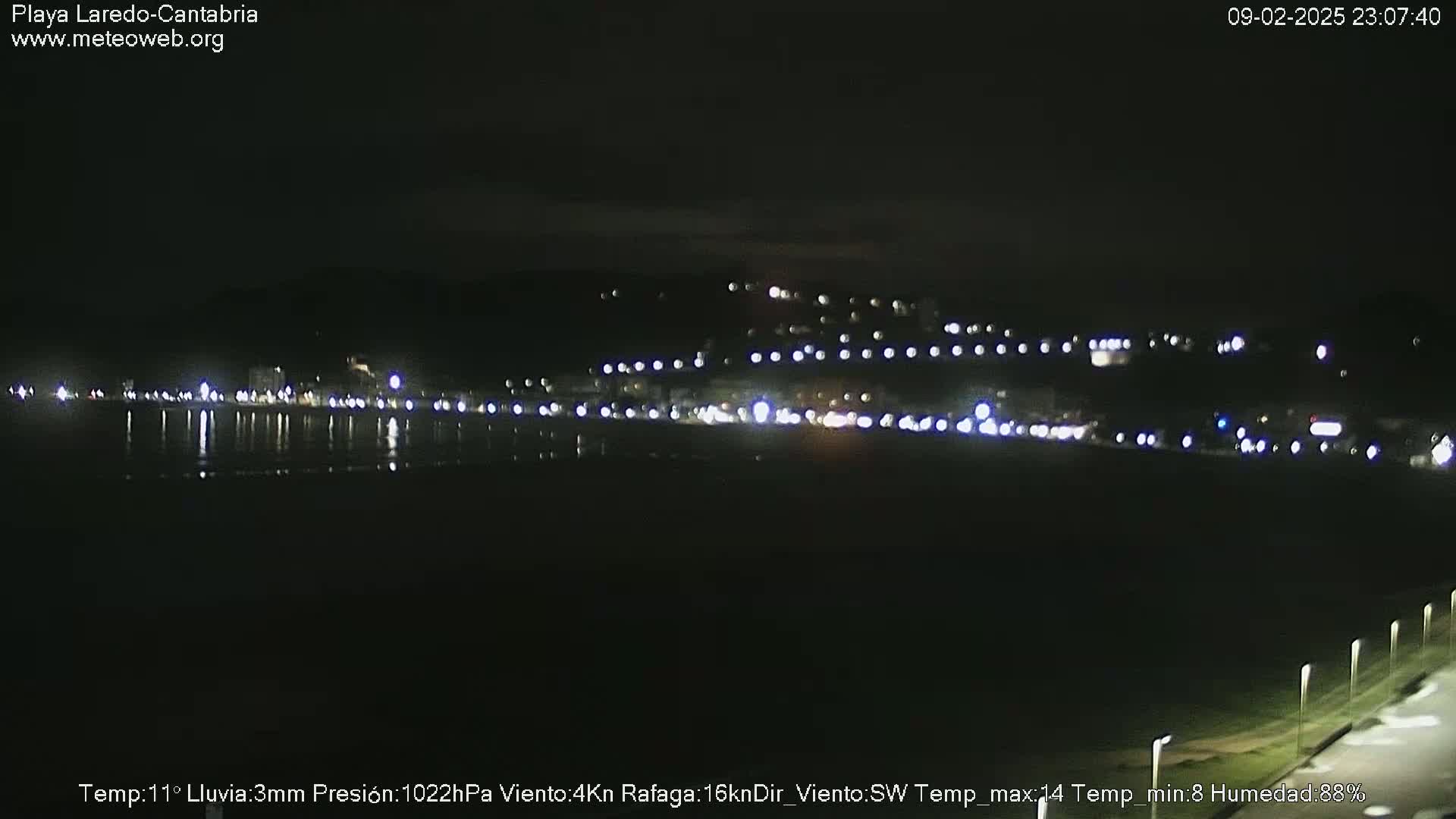A nighttime view of a coastal town with its lights reflected on dark water under a slightly overcast sky with light rain.
