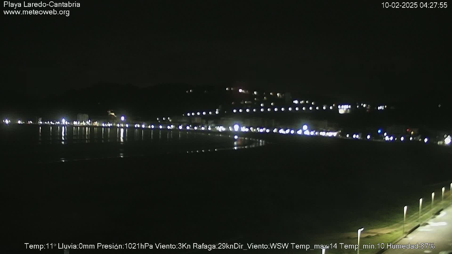 A dark, nighttime beach scene shows a line of lights reflecting in calm water, with a hill dotted with lights in the background.