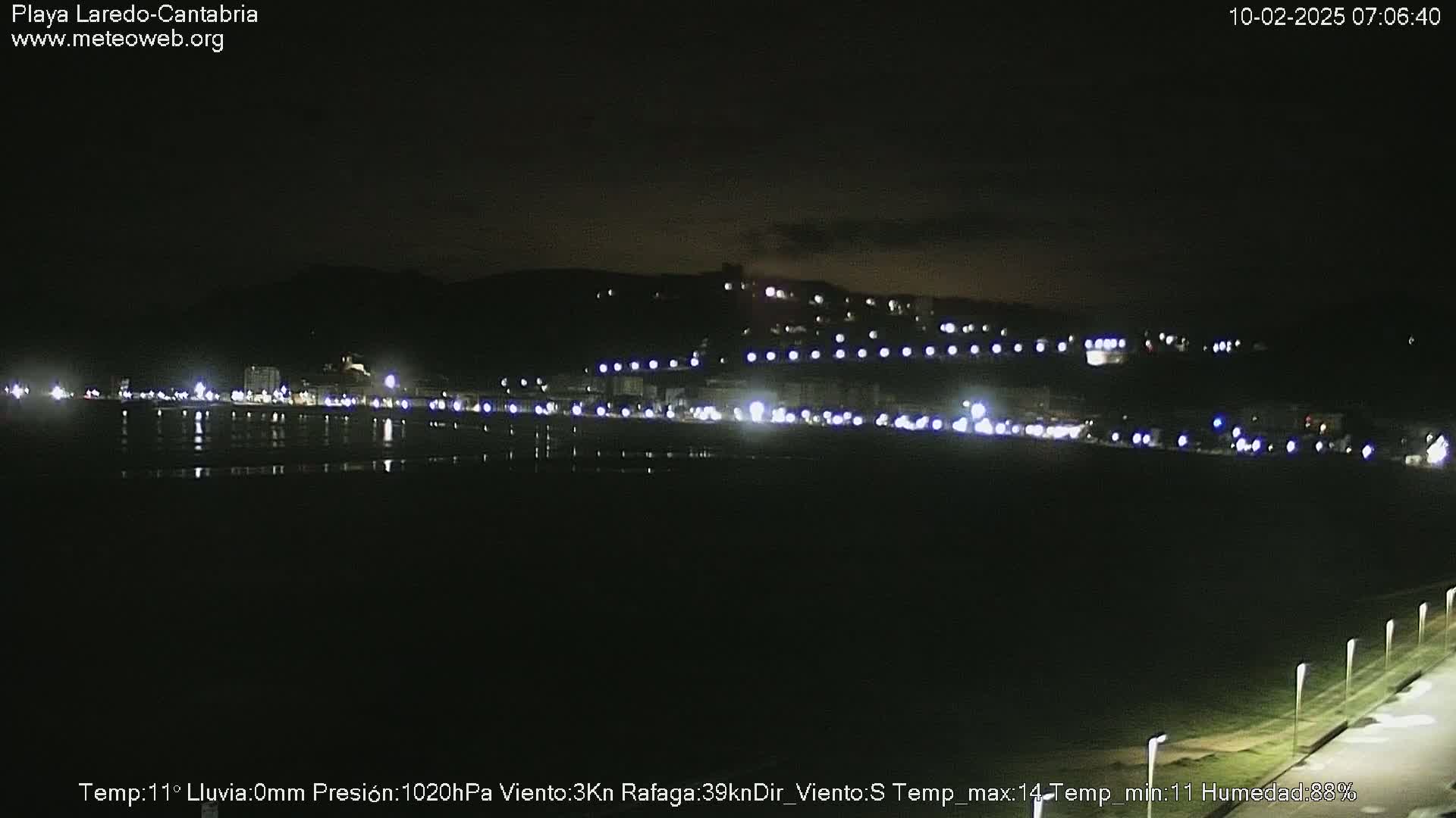 A nighttime view of a dark, calm beach with a town illuminated by lights on a hill in the background.