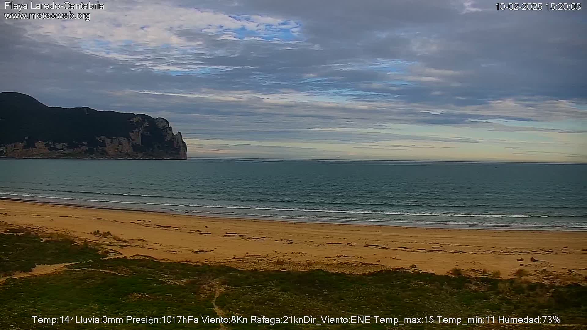 A sandy beach stretches to a calm sea under a mostly cloudy sky, with a dark, rocky headland visible in the distance.