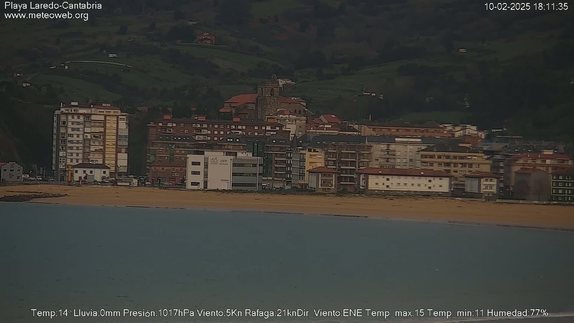 A coastal town with multi-story buildings and a sandy beach is seen from across calm water under overcast skies.