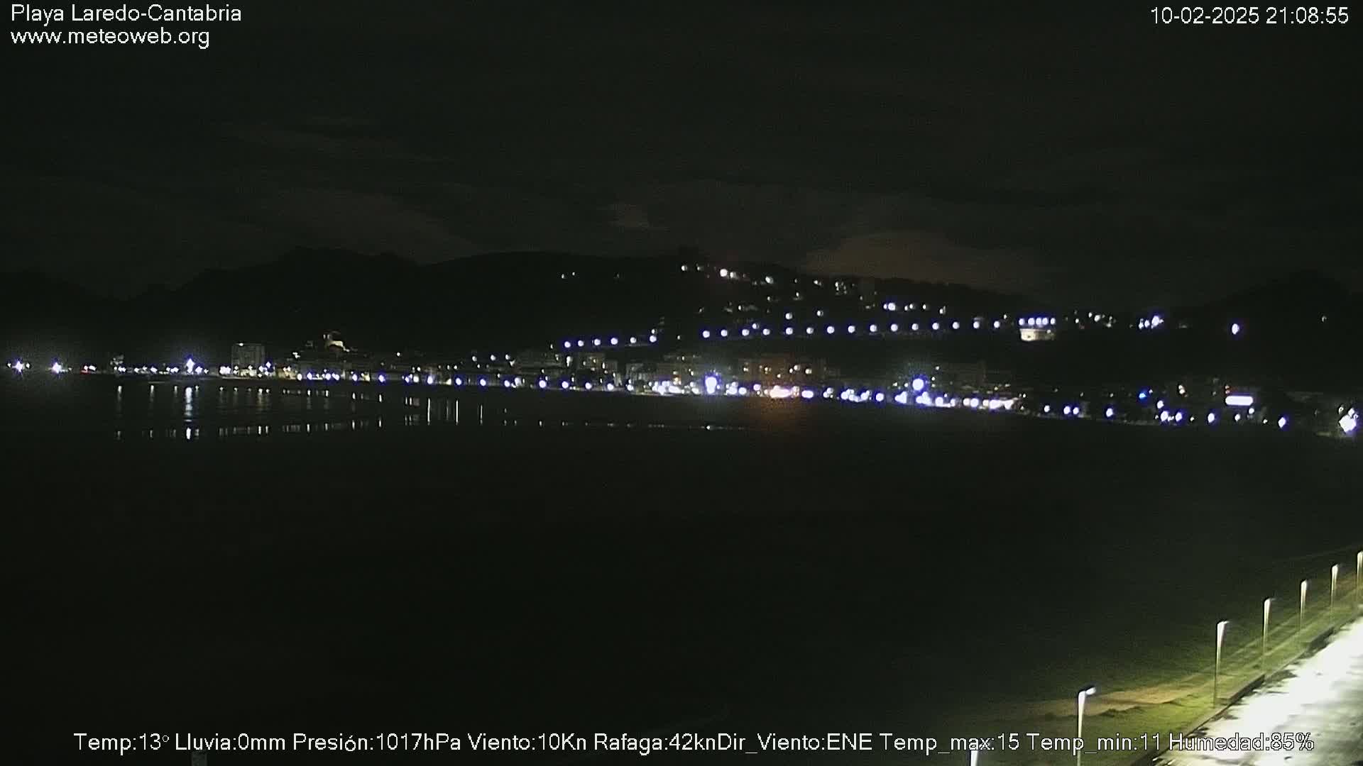 A nighttime view of a dark, calm beach with a town illuminated by lights on a hill in the background.