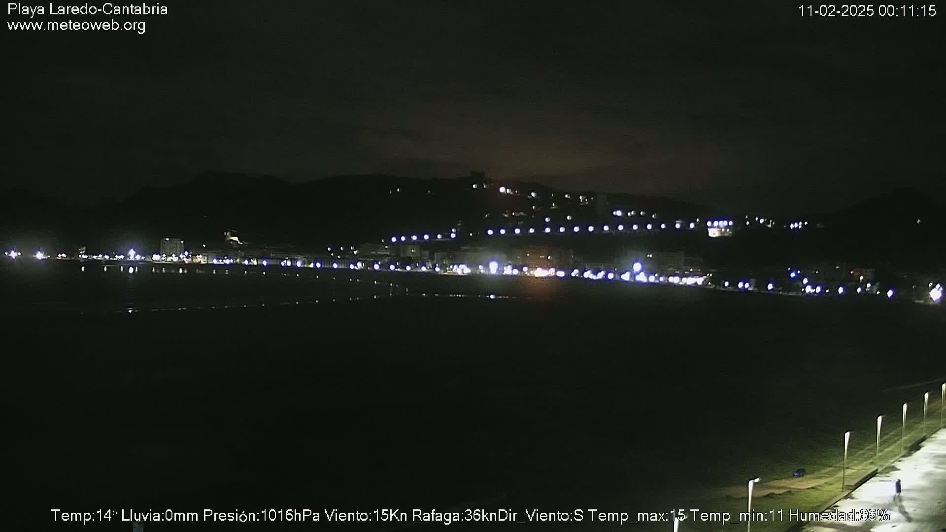 A nighttime view of a dark, seemingly empty beach with a line of lights from buildings and streetlights on the hill in the background.