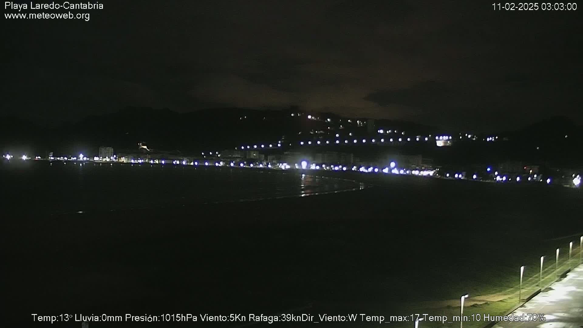 A dark nighttime view of a coastline with a town illuminated by lights in the distance, under a mostly clear sky.