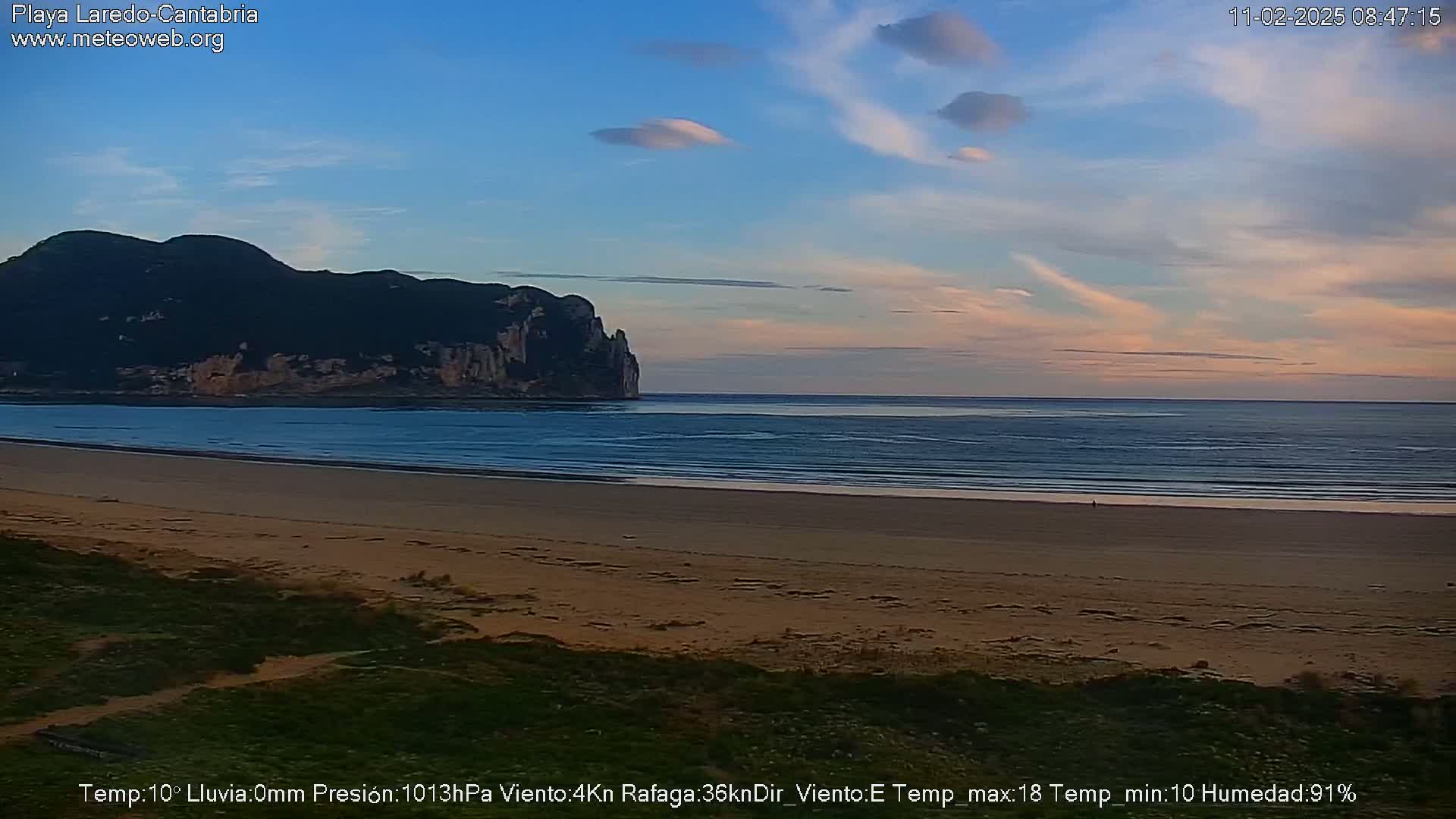 A calm ocean beach at sunset, with a dark, rocky headland in the background under a mostly clear sky.