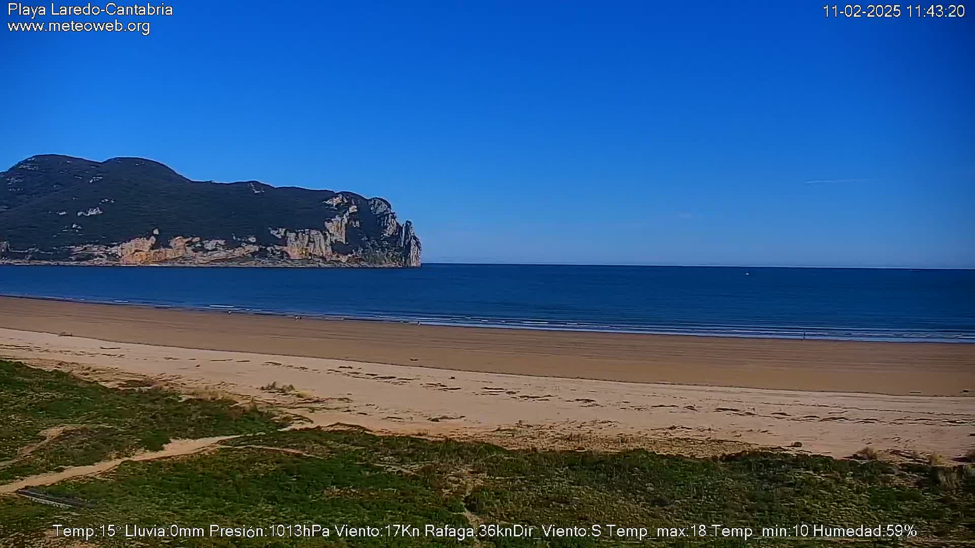 A sandy beach stretches towards a calm ocean under a clear blue sky, with a large, dark-green hill rising from the water in the background.