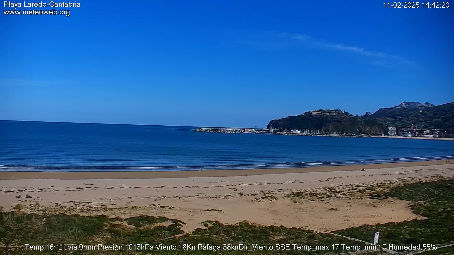 A sandy beach stretches along a calm ocean under a clear blue sky, with hills and a small town visible in the distance.