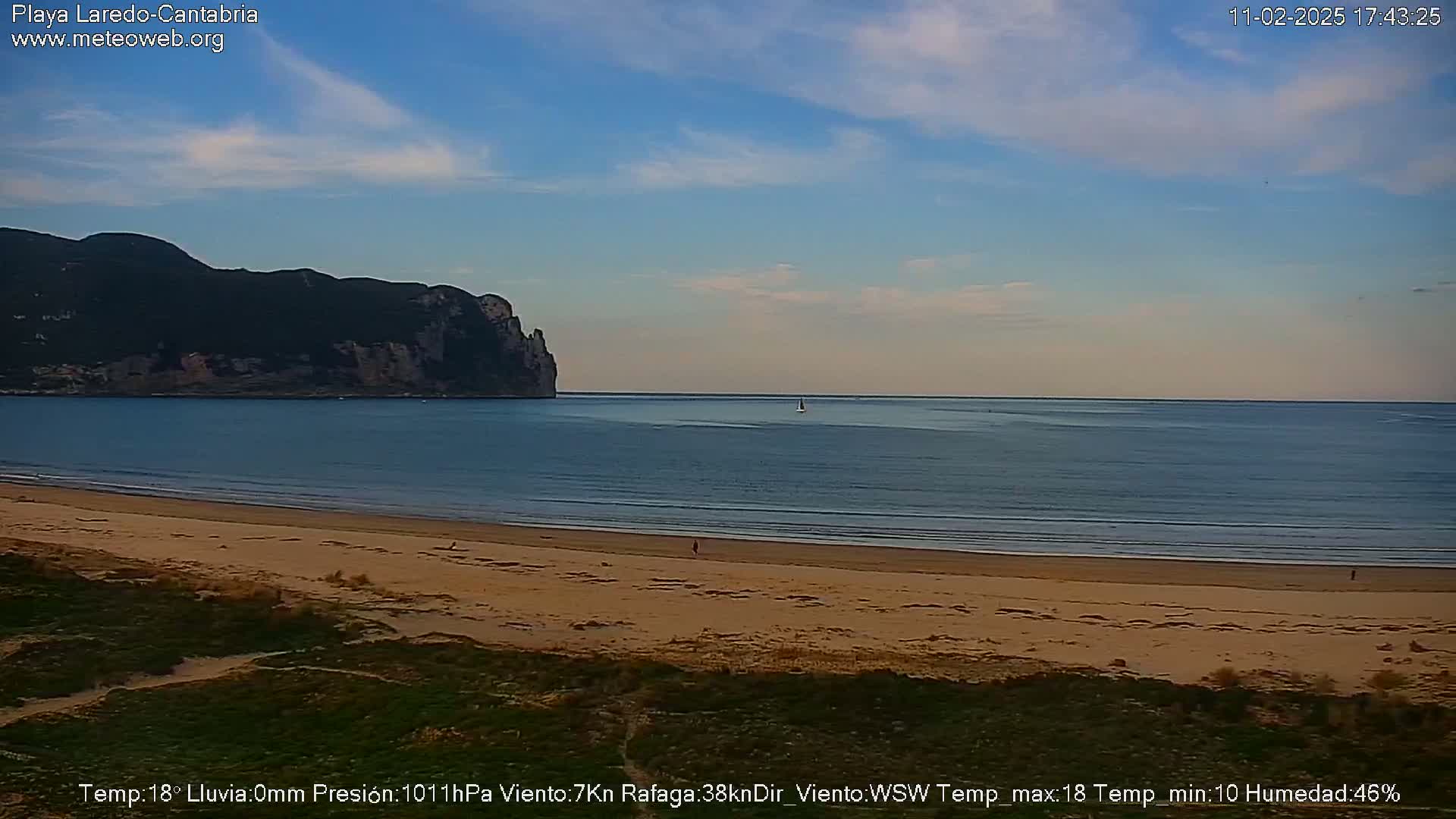 A sandy beach stretches to a calm ocean under a mostly clear sky, with a dark mountain visible in the distance.