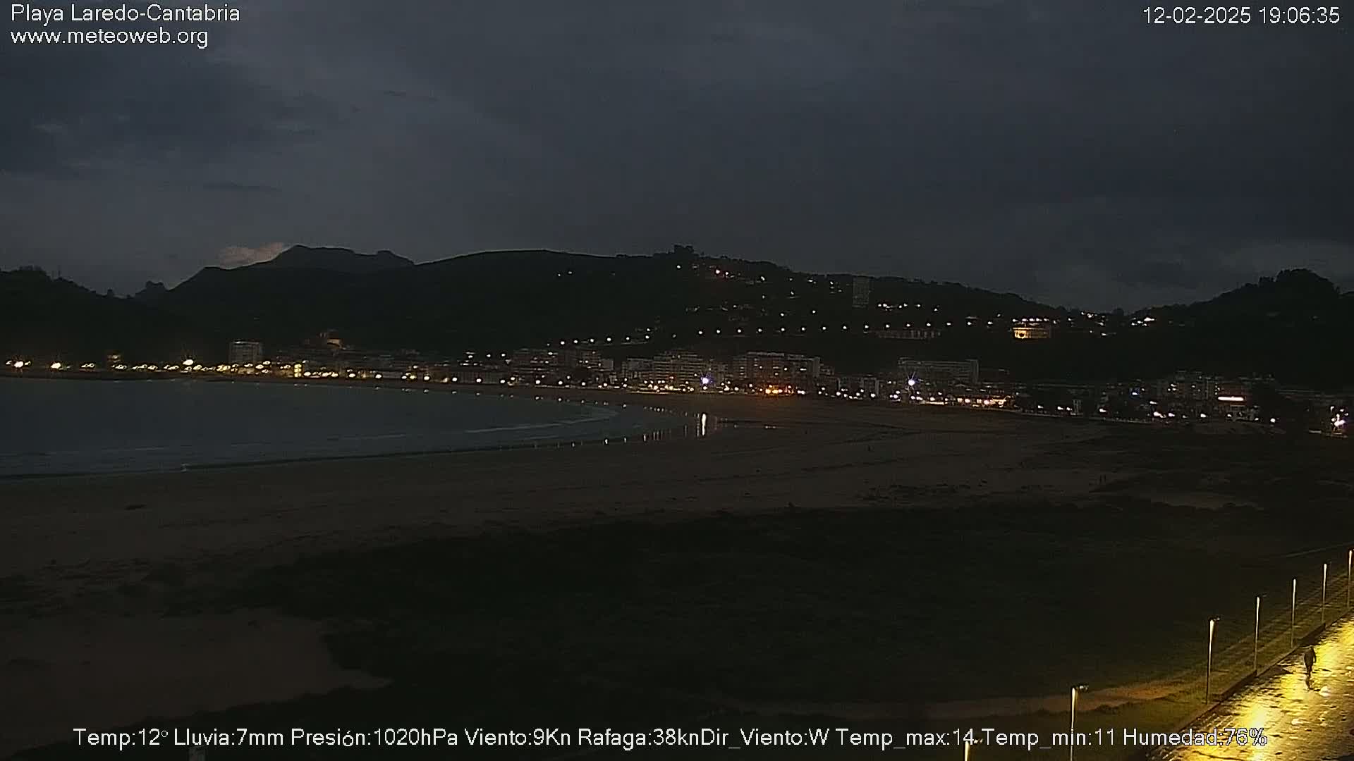 A dark, nighttime view of a beach with a town illuminated on a hill in the background under a cloudy sky.