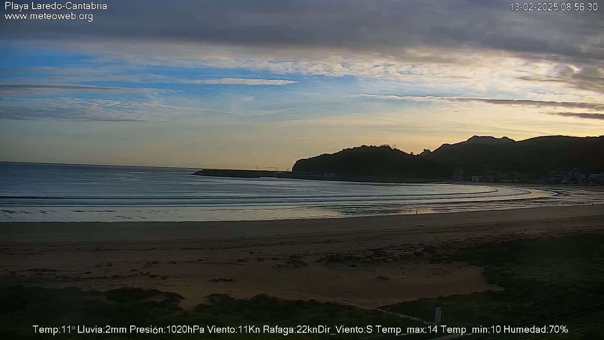 A sandy beach curves gently around a calm bay, backed by a dark hill under a mostly clear, twilight sky.