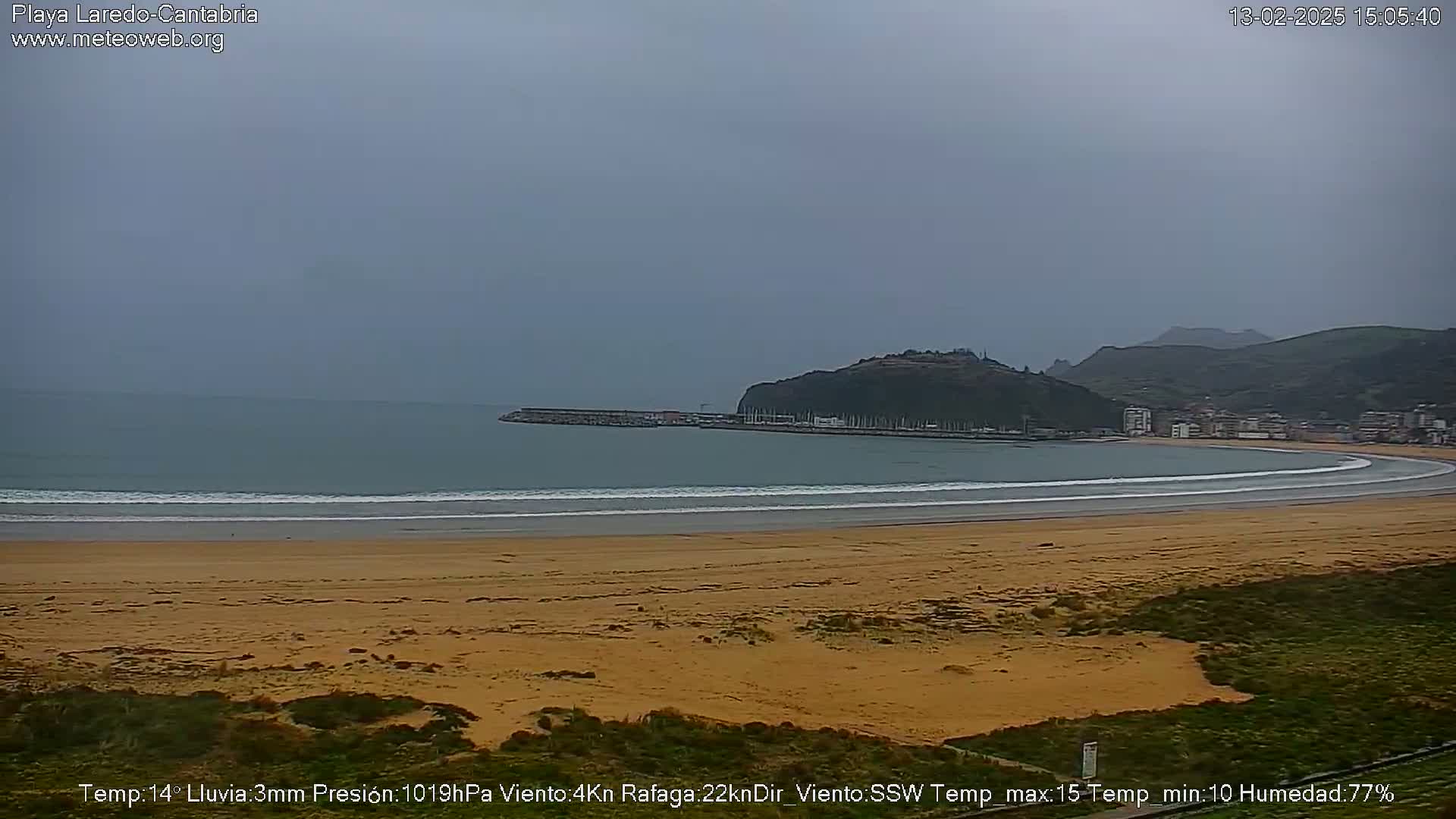 A sandy beach curves gently around a bay under an overcast sky, with small waves lapping the shore and a town visible in the distance.