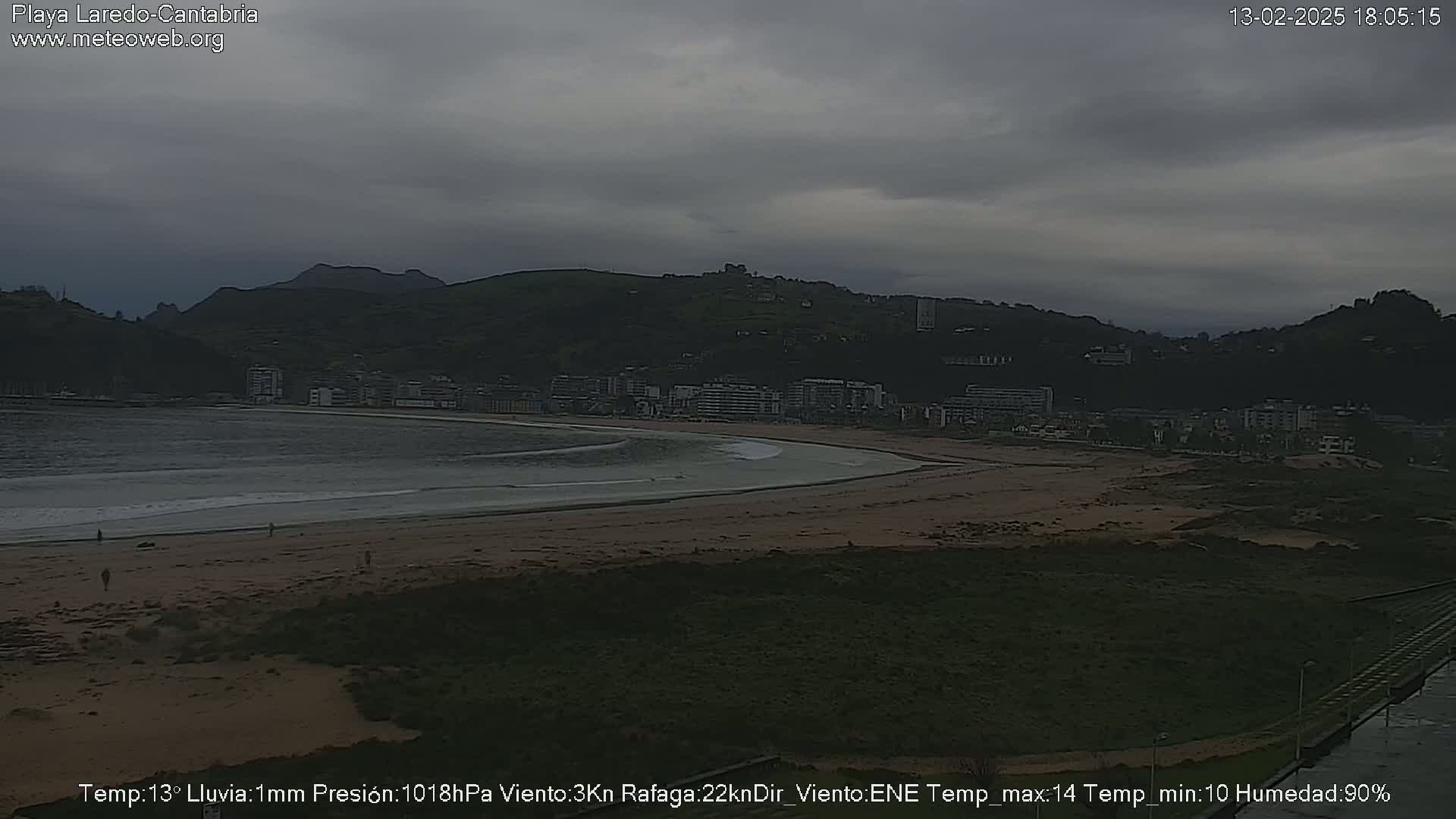 A beach with a town and hills in the background under an overcast sky.
