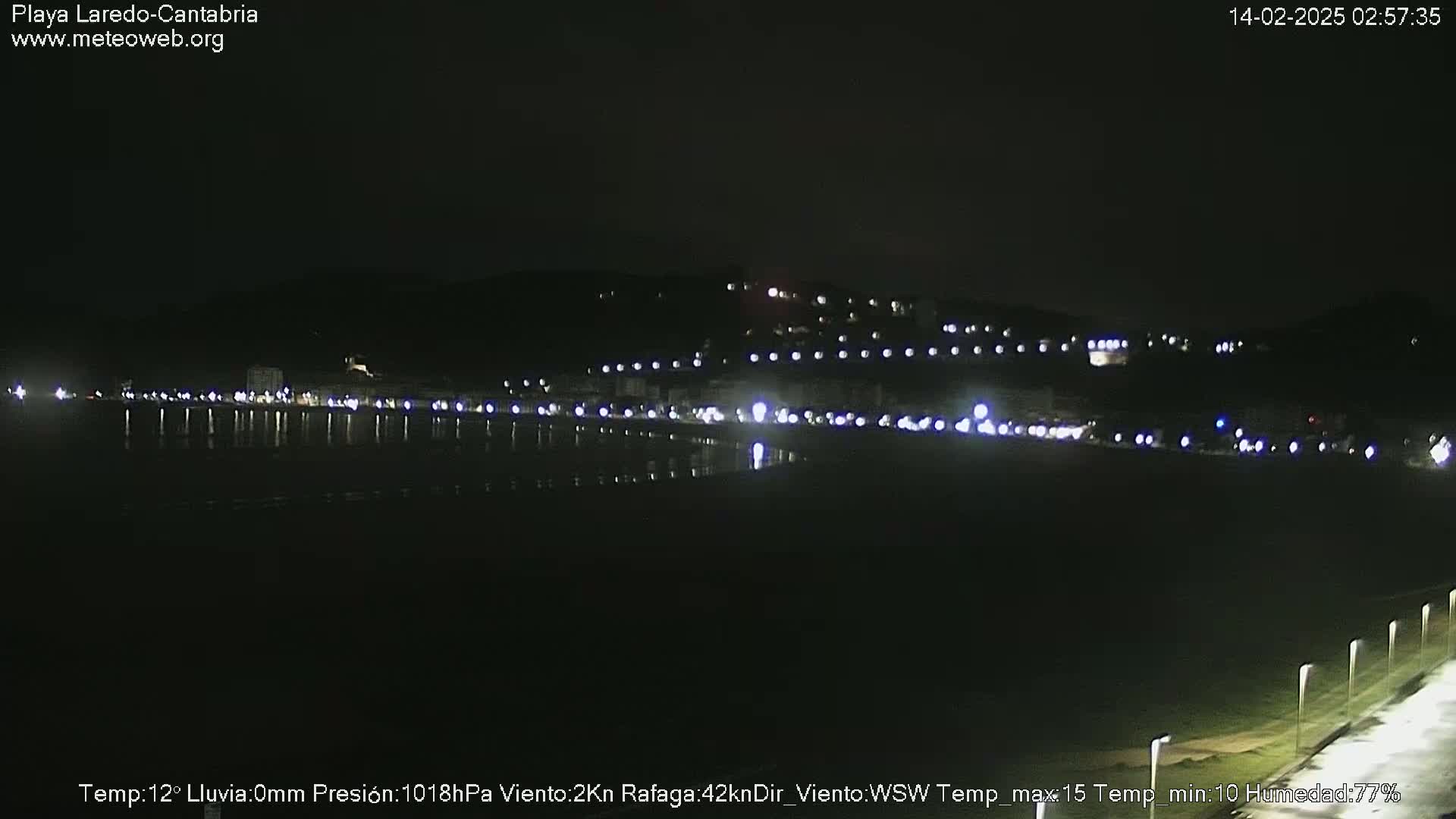 A nighttime view of a dark, calm body of water reflecting the lights of a coastal town nestled against a hillside.