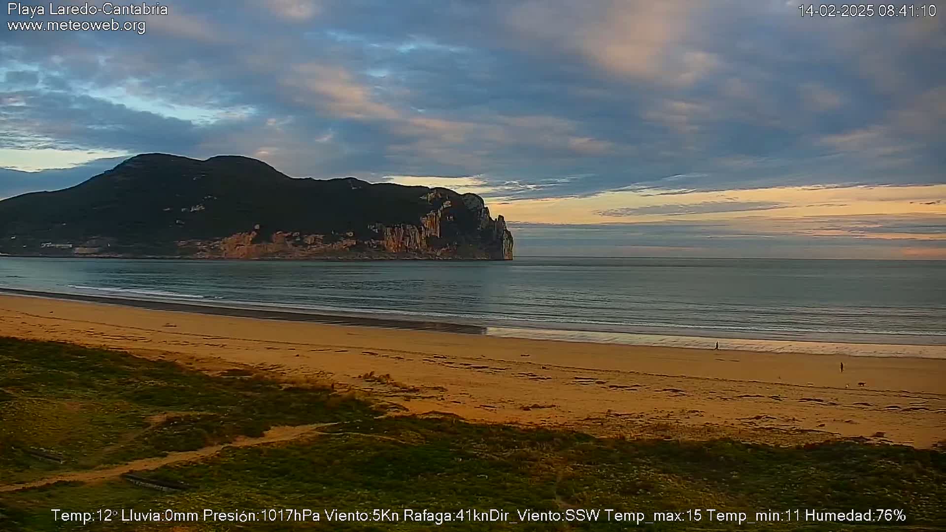 A sandy beach stretches towards a calm ocean under a partly cloudy sky, with a large dark mountain visible in the distance.