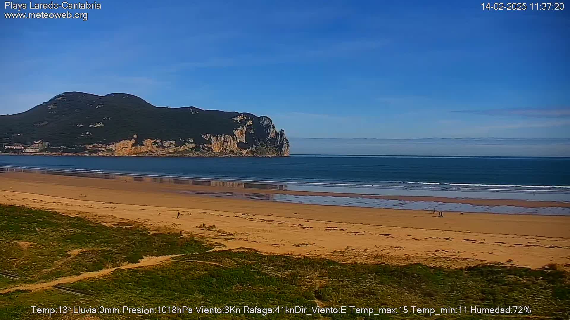 A sandy beach stretches out to a calm ocean under a clear blue sky, with a dark green hill rising in the background.