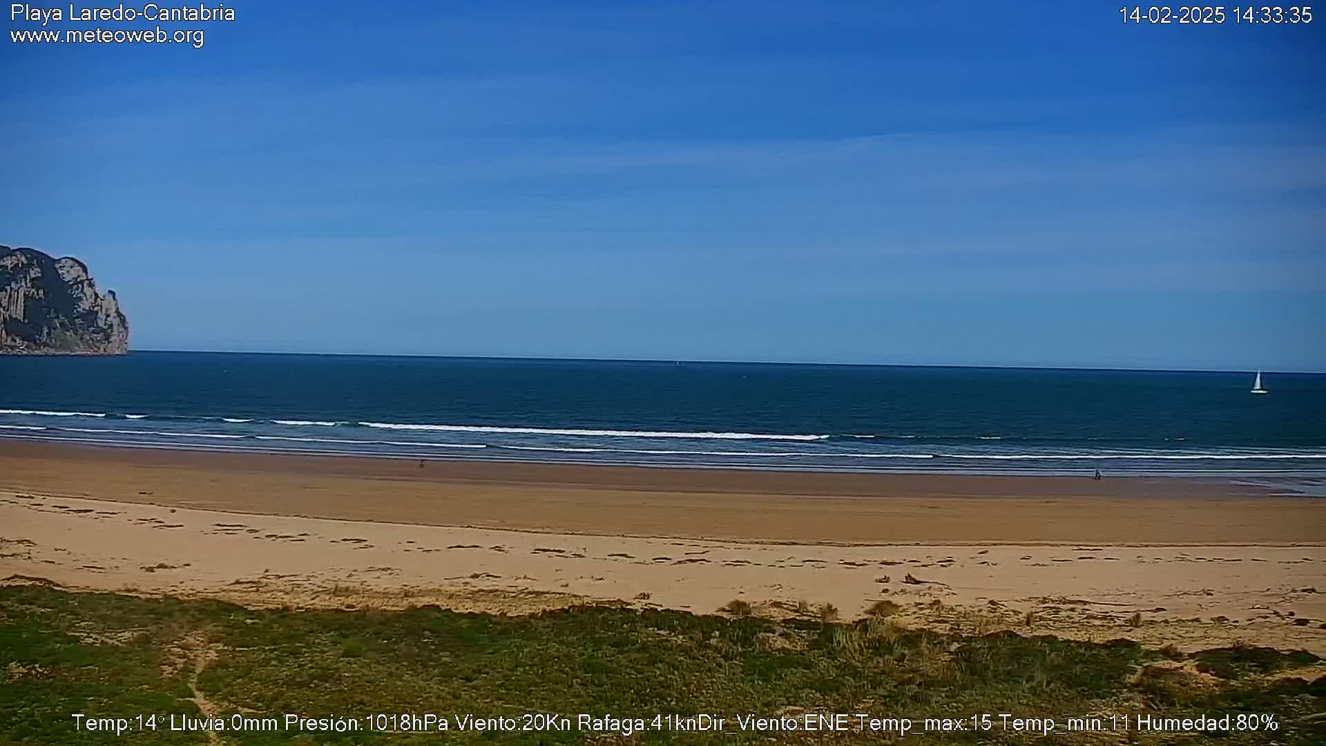 A sandy beach stretches towards a calm ocean under a clear blue sky, with a distant cliff visible to the left and a lone sailboat on the horizon.