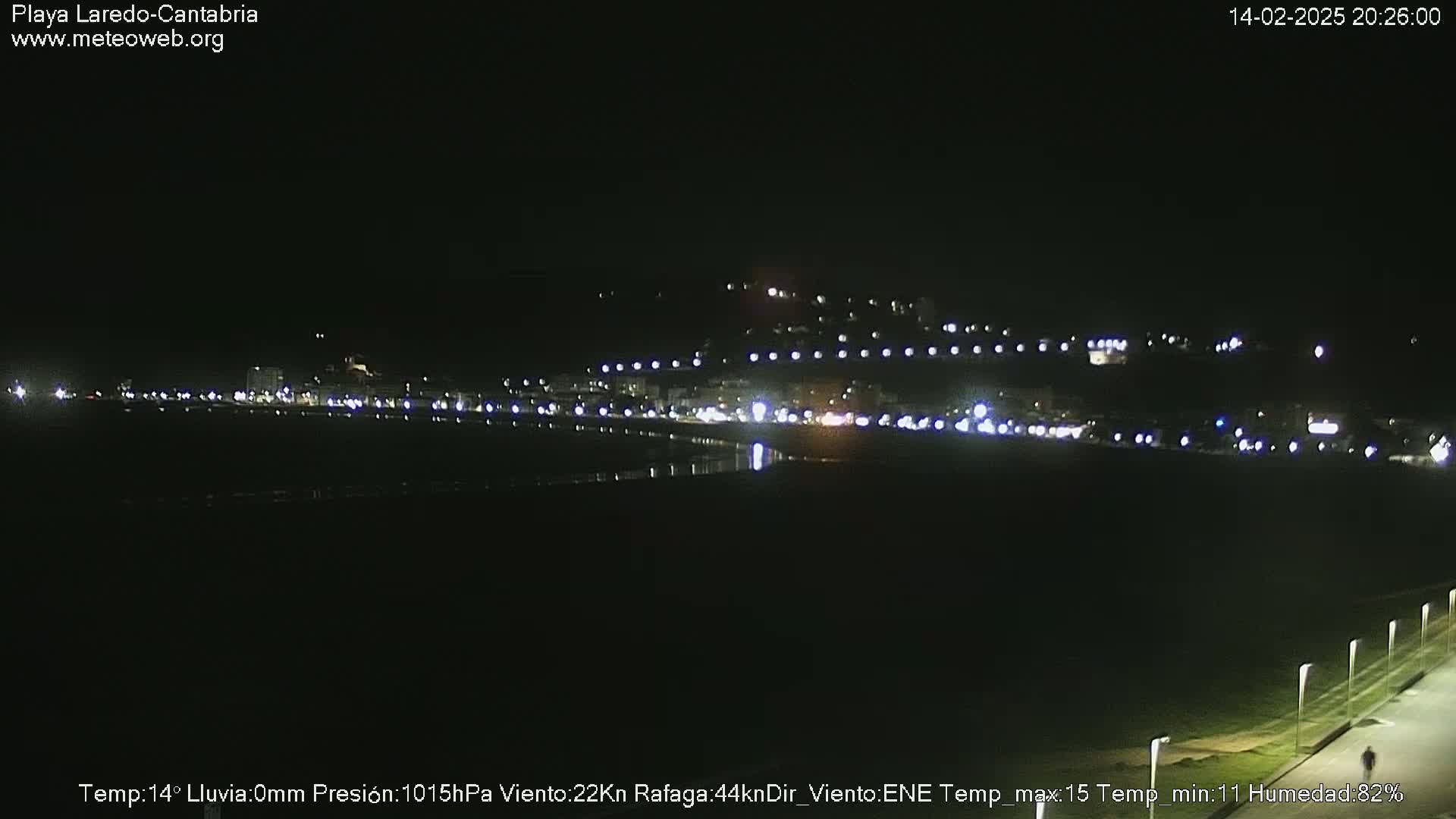 A nighttime long shot of a coastal town with its lights reflecting on dark water under a clear, dark sky.