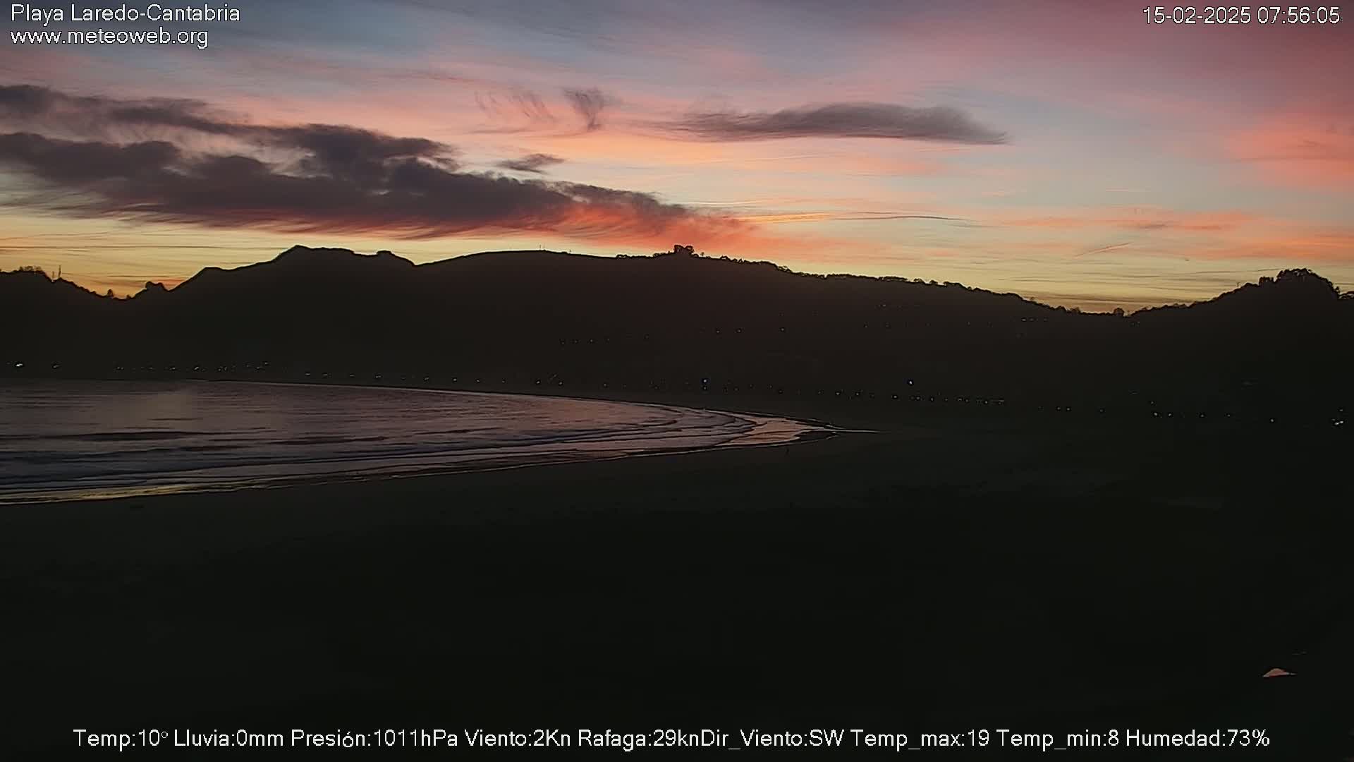 A dark, sandy beach stretches beneath a twilight sky displaying soft pink and orange hues above silhouetted hills.