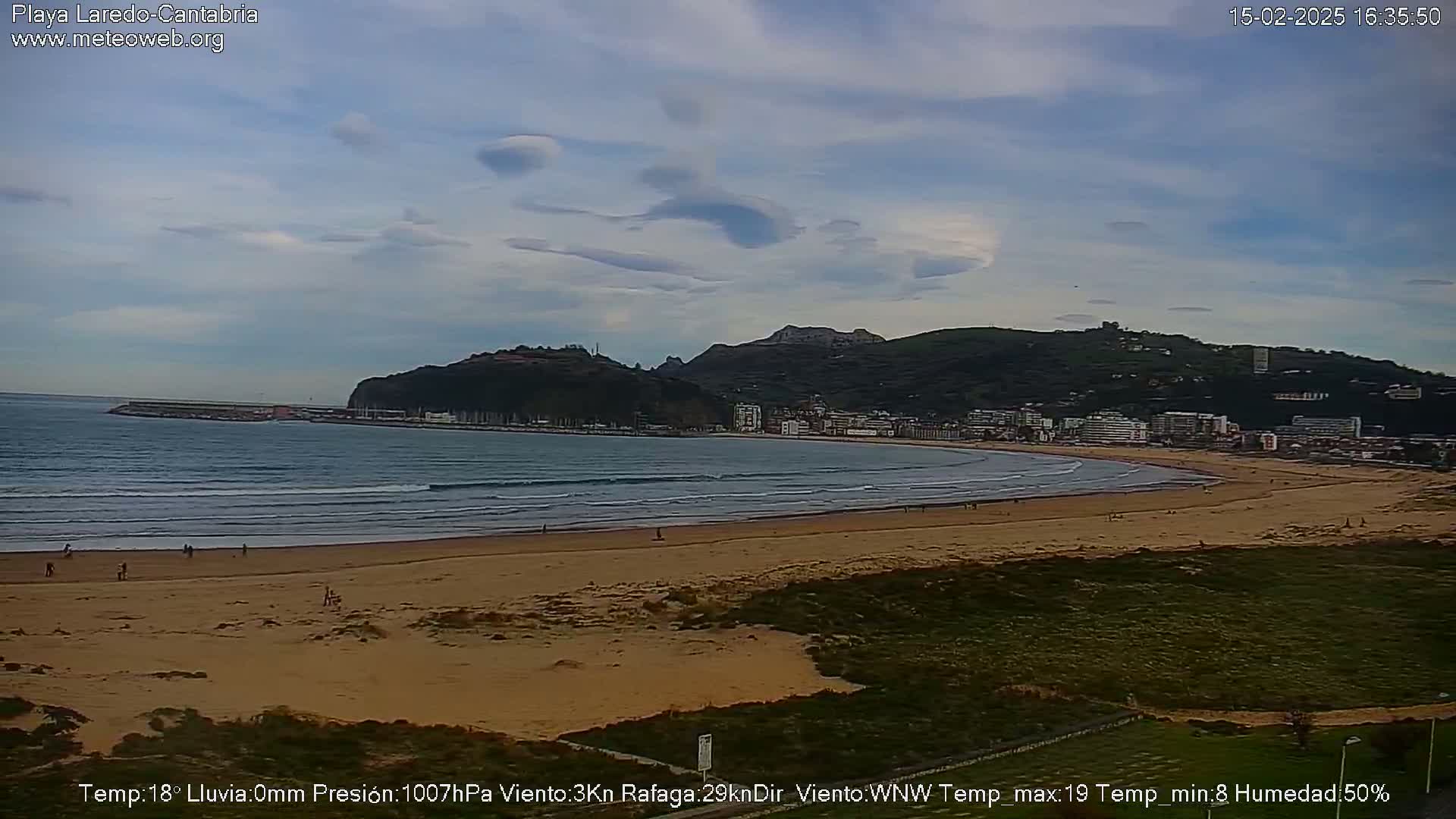 A sandy beach with gentle waves laps the shore, under a partly cloudy sky, with a town and hills in the background.