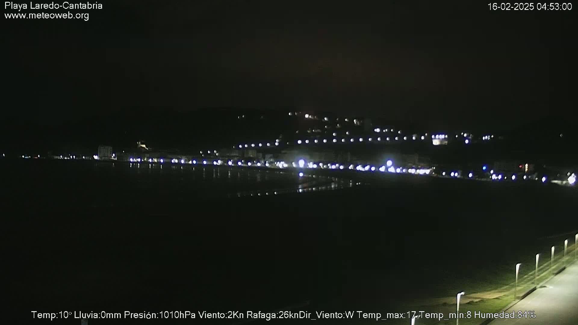 A nighttime view of a dark, flat coastal area with a line of lights along the horizon and a few lights visible on a hill in the distance.