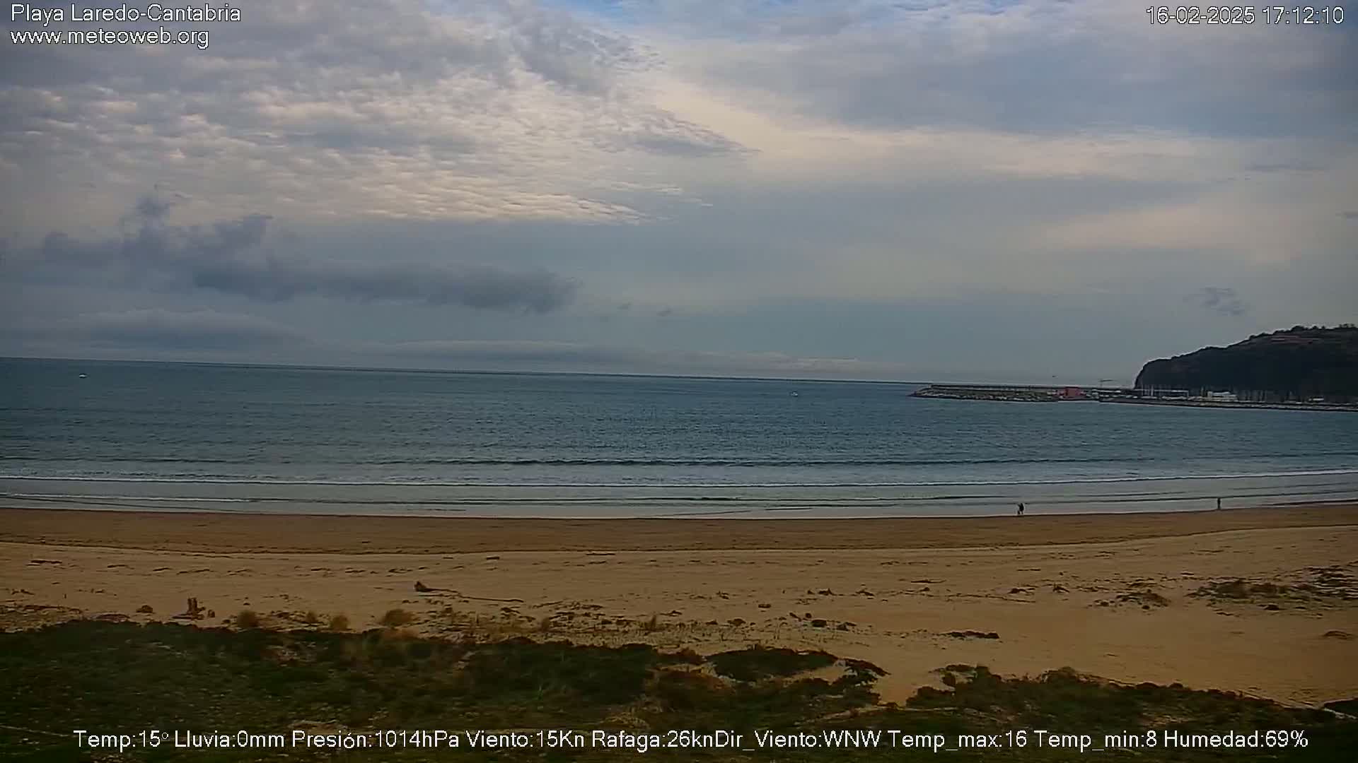 A sandy beach stretches to a calm ocean under a mostly cloudy sky.