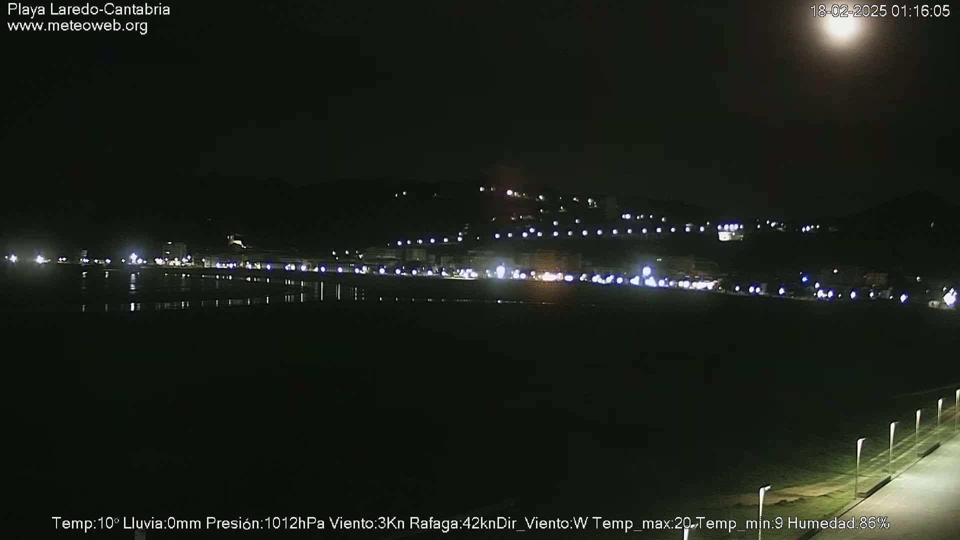 A nighttime view of a dark, flat coastal area with a line of lights on the horizon under a mostly clear, moonlit sky.