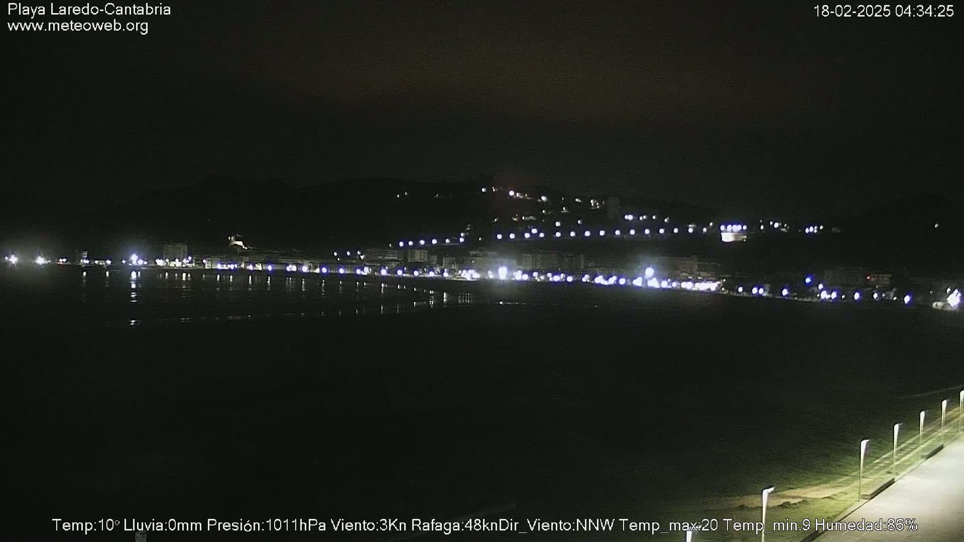 A nighttime view of a dark, seemingly calm beach with a line of lights illuminating buildings on a hill in the background.