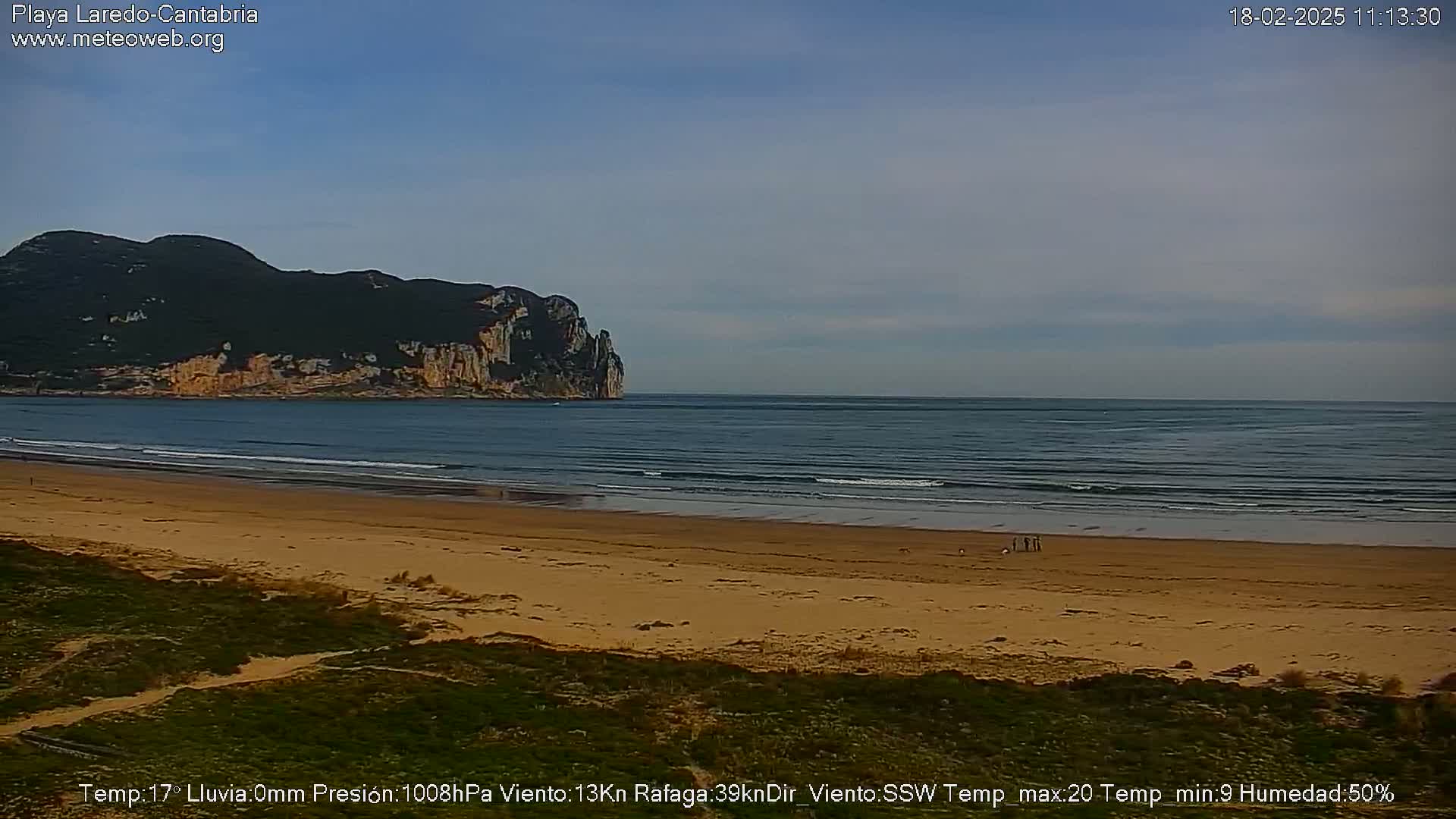 A sandy beach stretches towards a calm ocean under a clear sky, with a large, dark-green hill visible in the distance.