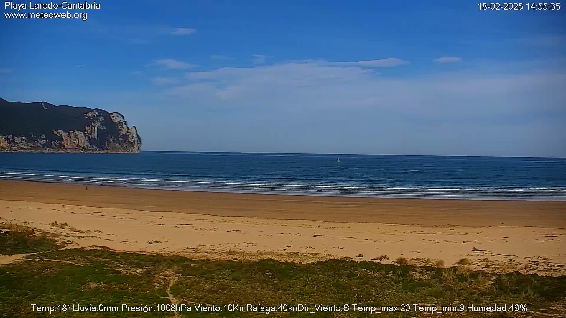 A sandy beach stretches to a calm ocean under a mostly clear blue sky, with a distant rocky cliff visible on the left.