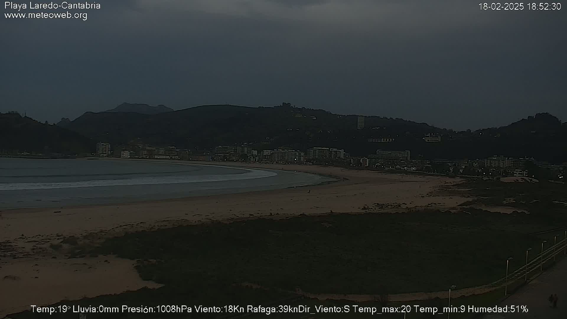 A dark, overcast beach scene shows a calm bay with a sandy shoreline and a town nestled against a hill in the background.