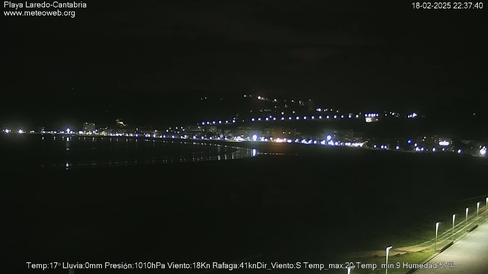 A nighttime coastal view shows a dark beach and a line of lights from buildings on a hill across the water.
