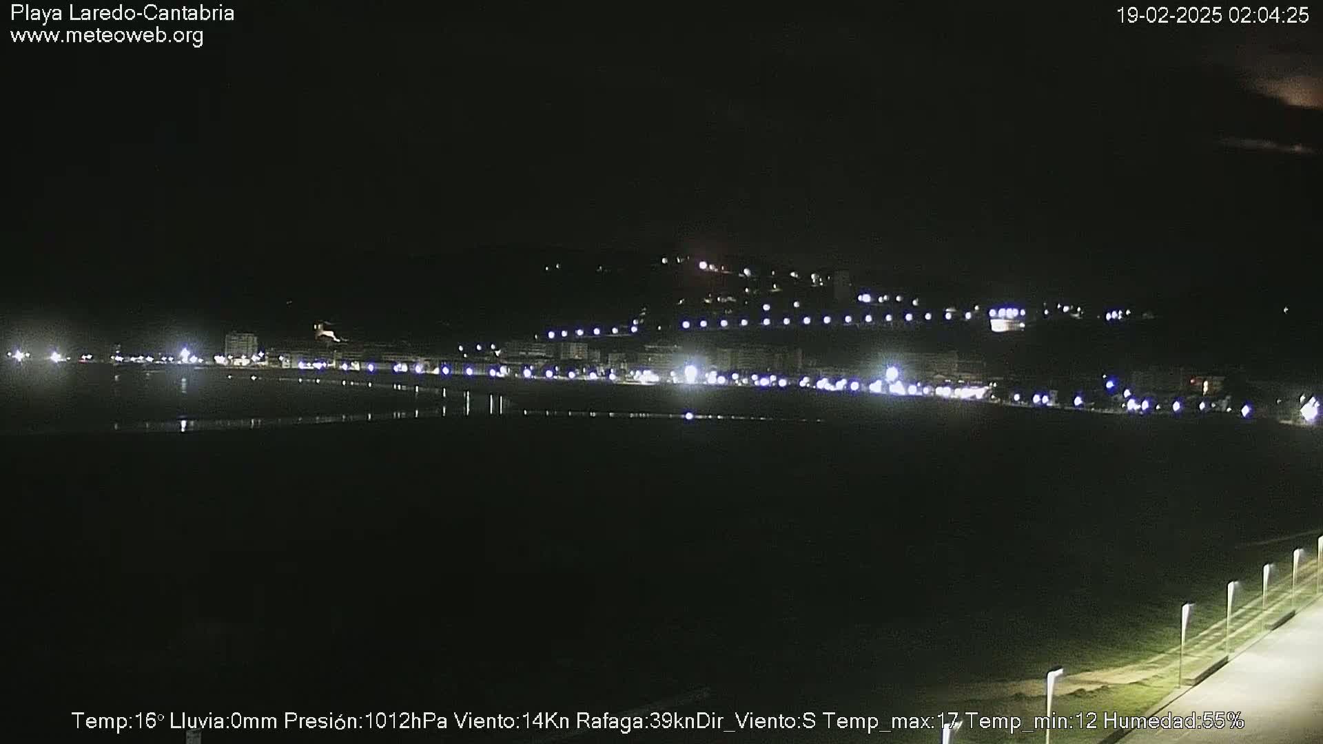 A nighttime view of a dark, flat coastal area with a line of lights from buildings on a hill in the background, under clear skies.