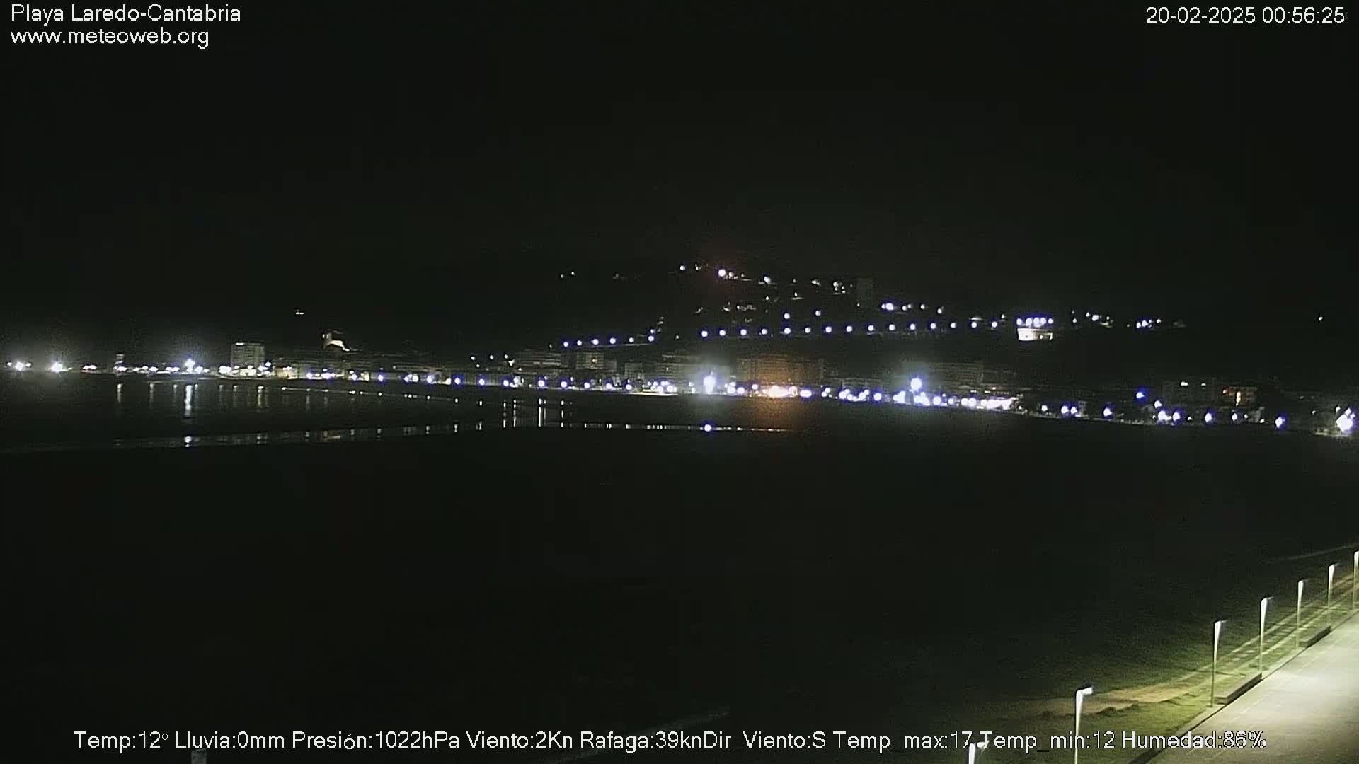 A nighttime view of a coastal town with lights illuminating the buildings and a dark, calm body of water reflecting the lights.