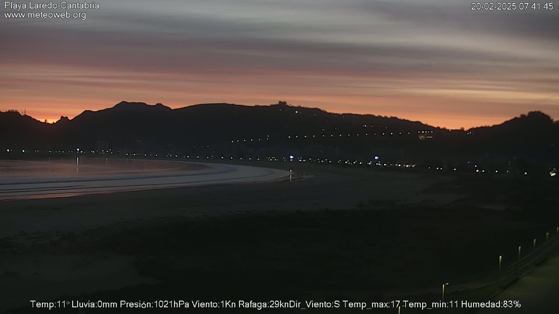 A dark beach at sunset, with a town and hills in the background under a mostly clear sky.
