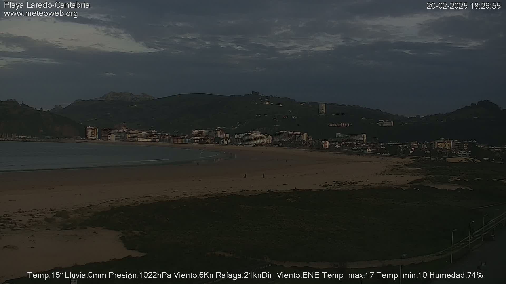 A beach with a town and dark mountains in the background under an overcast sky.
