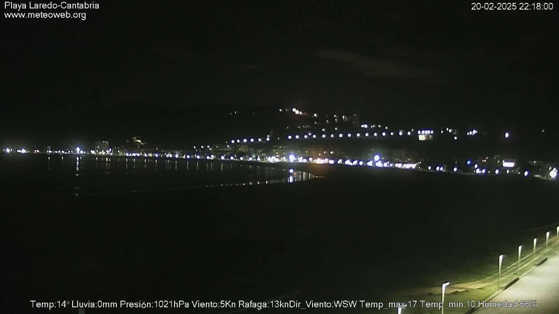 A nighttime view of a dimly lit coastal town with a dark beach and calm water, under a clear sky.