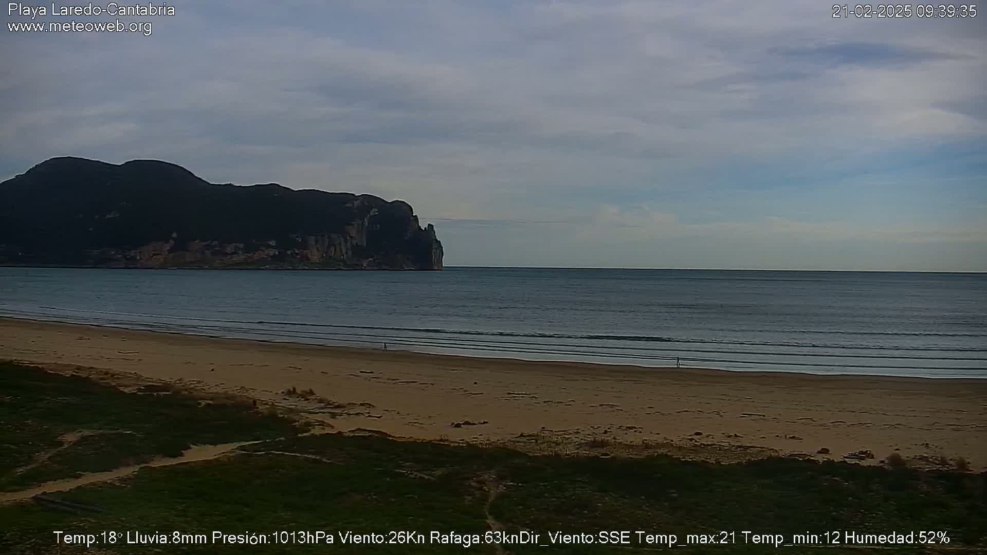 A sandy beach stretches to a calm sea under an overcast sky, with a dark mountain visible in the distance.
