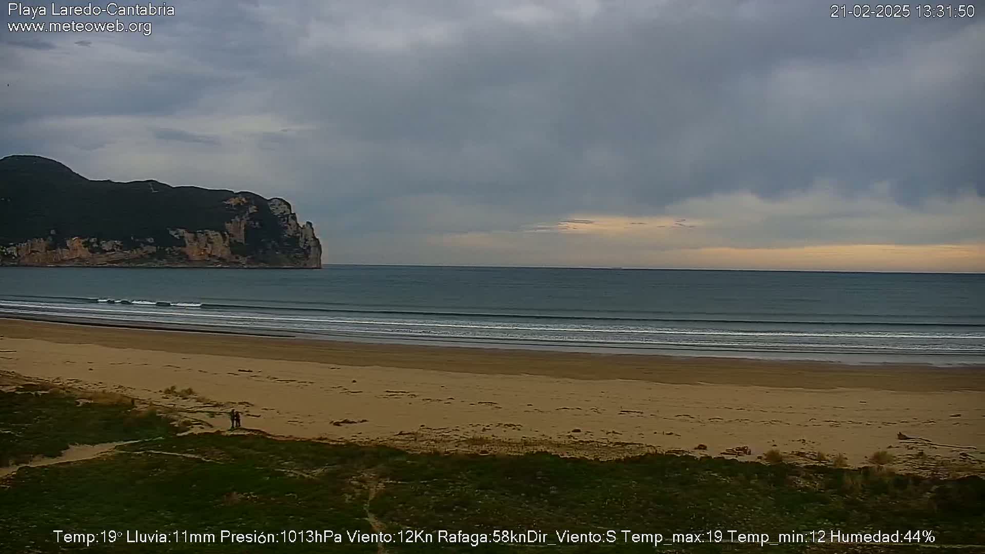 A sandy beach with gentle waves stretches toward a rocky cliff under a mostly cloudy sky.